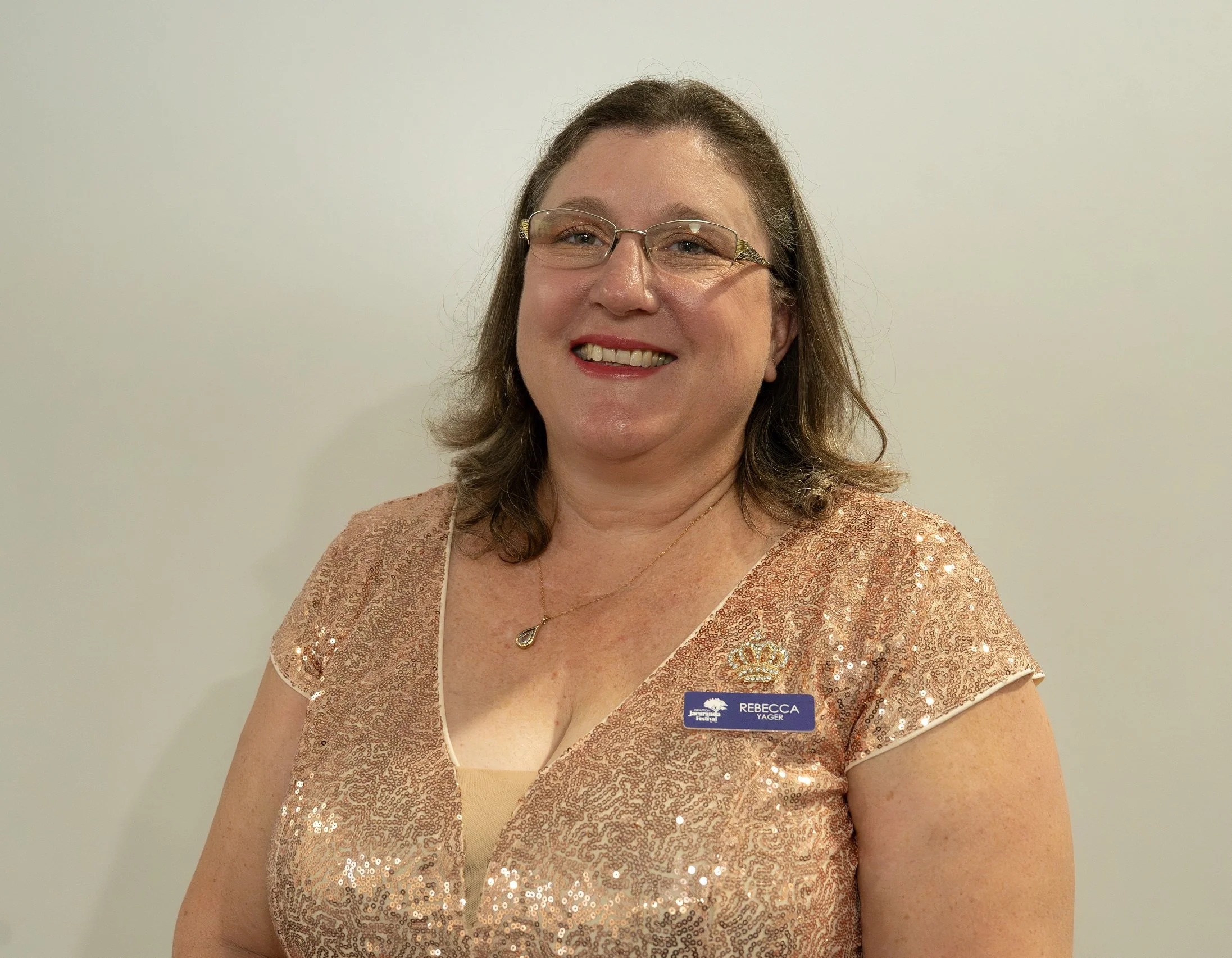 Professional woman with glasses, smiling, wearing a rose gold sequined dress, a name tag that reads 'Rebecca Yager' and a small crown pin, standing in front of a plain light-colored wall.