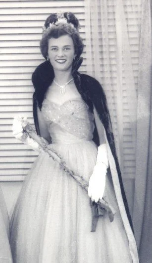 Black and white photo of a woman in a formal gown holding a prop, standing in front of curtains.