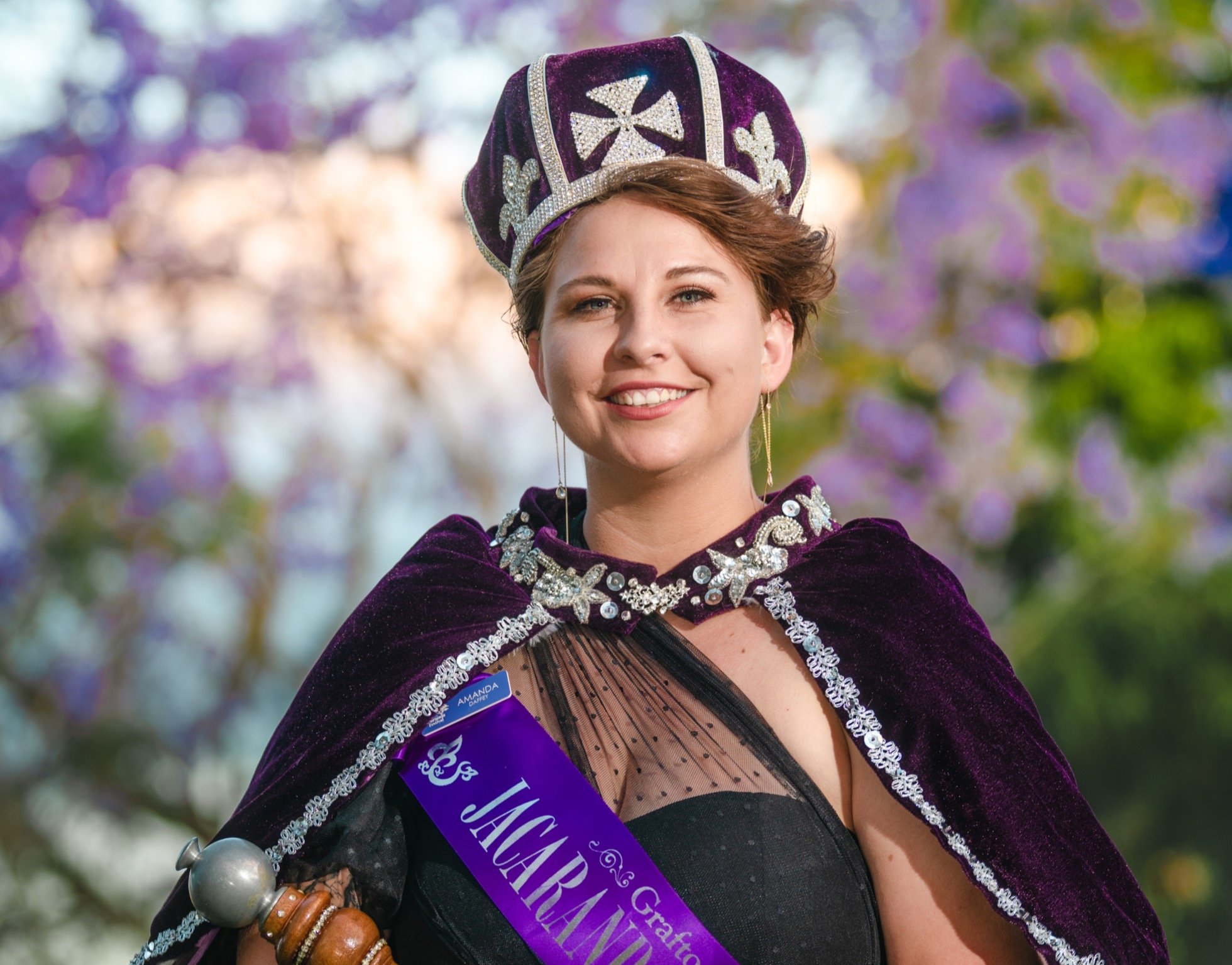 A woman wearing a purple crown-like hat with silver embellishments and a matching velvet cape, smiling outdoors with a colorful bokeh background. She has a purple sash with white text and is dressed in a black mesh top with polka dots. She accessorizes with long earrings, bracelets, and holds a silver ball on her wrist.