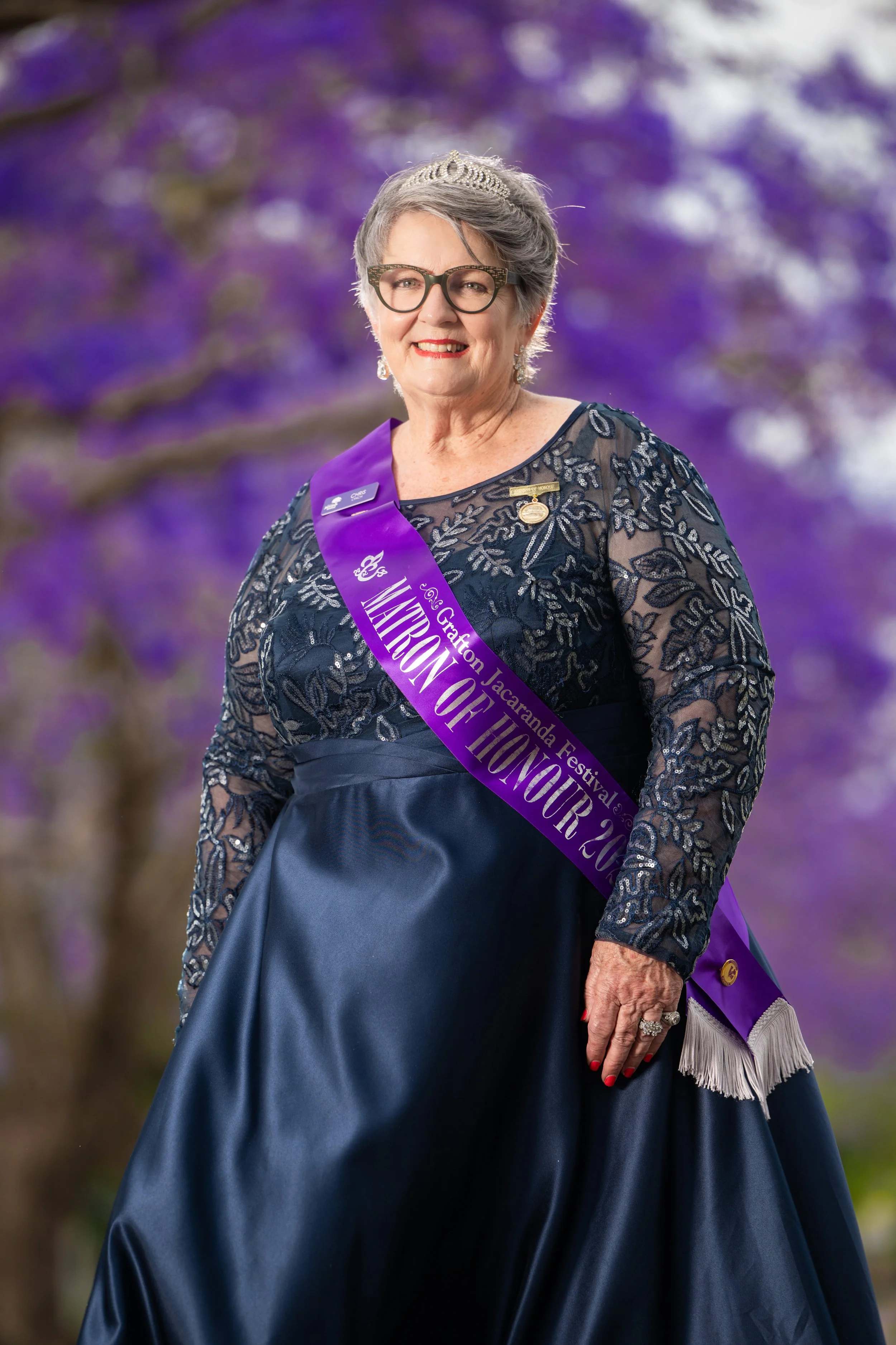 An elderly woman wearing glasses, a tiara, a dark blue dress with lace sleeves, and a purple sash. She is smiling and standing outdoors with a purple-flowered tree in the background.