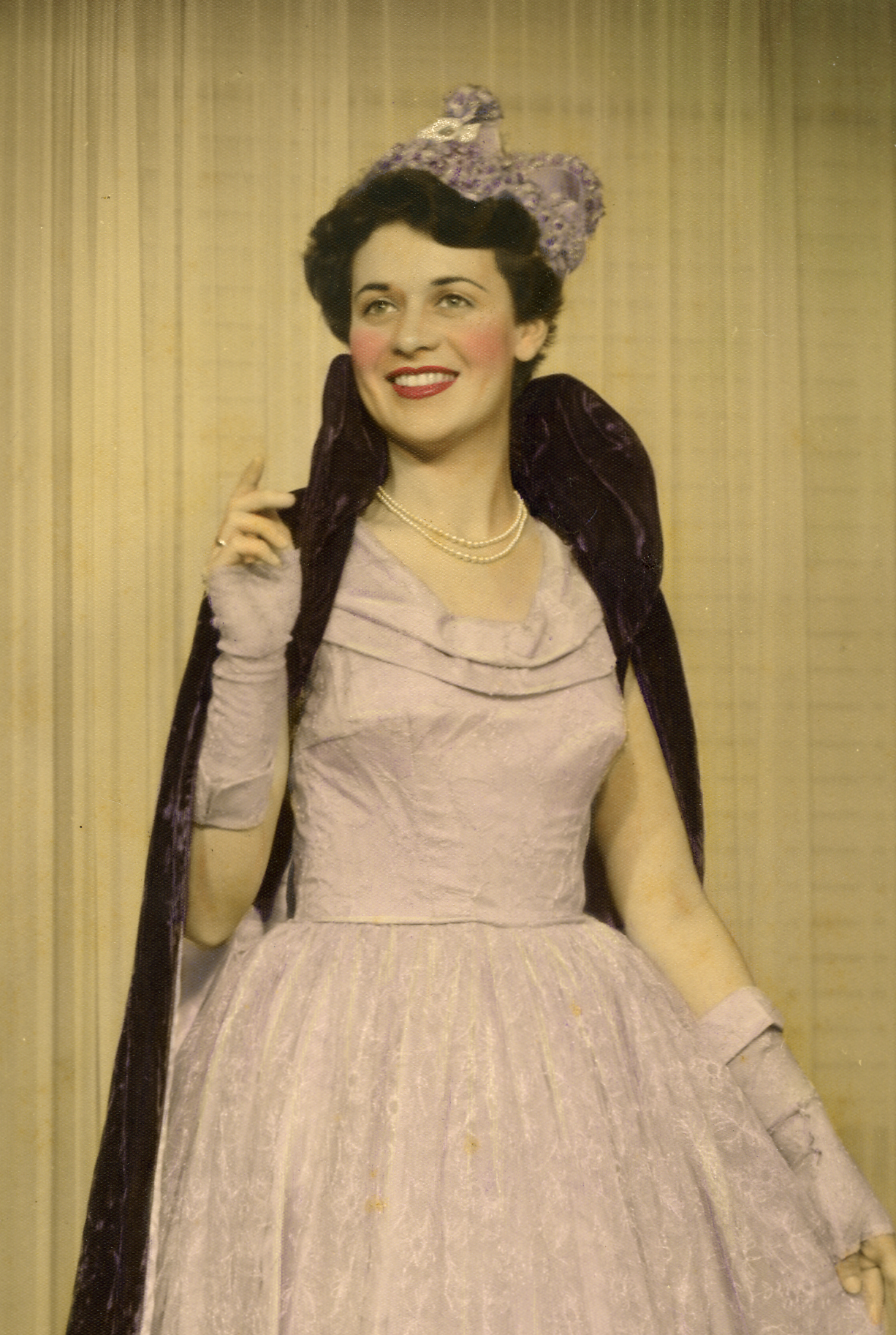 A woman dressed in vintage fashion, wearing a light-colored dress with a full skirt, pearl necklace, and a decorative headpiece, posing against a yellow striped background.