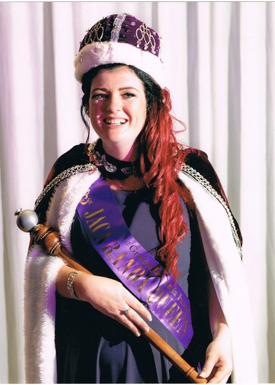 A woman with red hair wearing a crowning hat and cape, holding a scepter, and wearing a sash that says 'Jewel Princess.' She is smiling in front of a curtain backdrop.