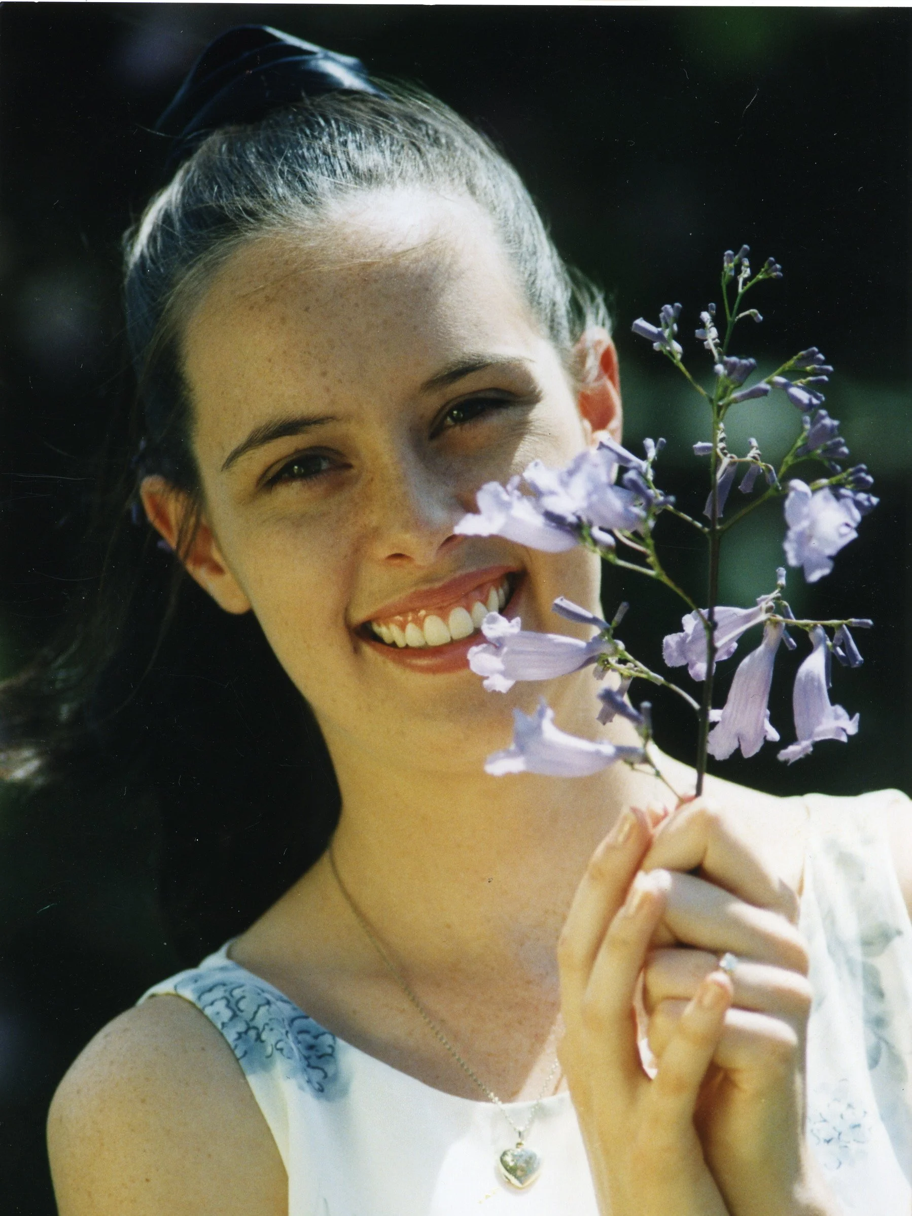 A smiling woman with dark hair holding a purple flower close to her face.