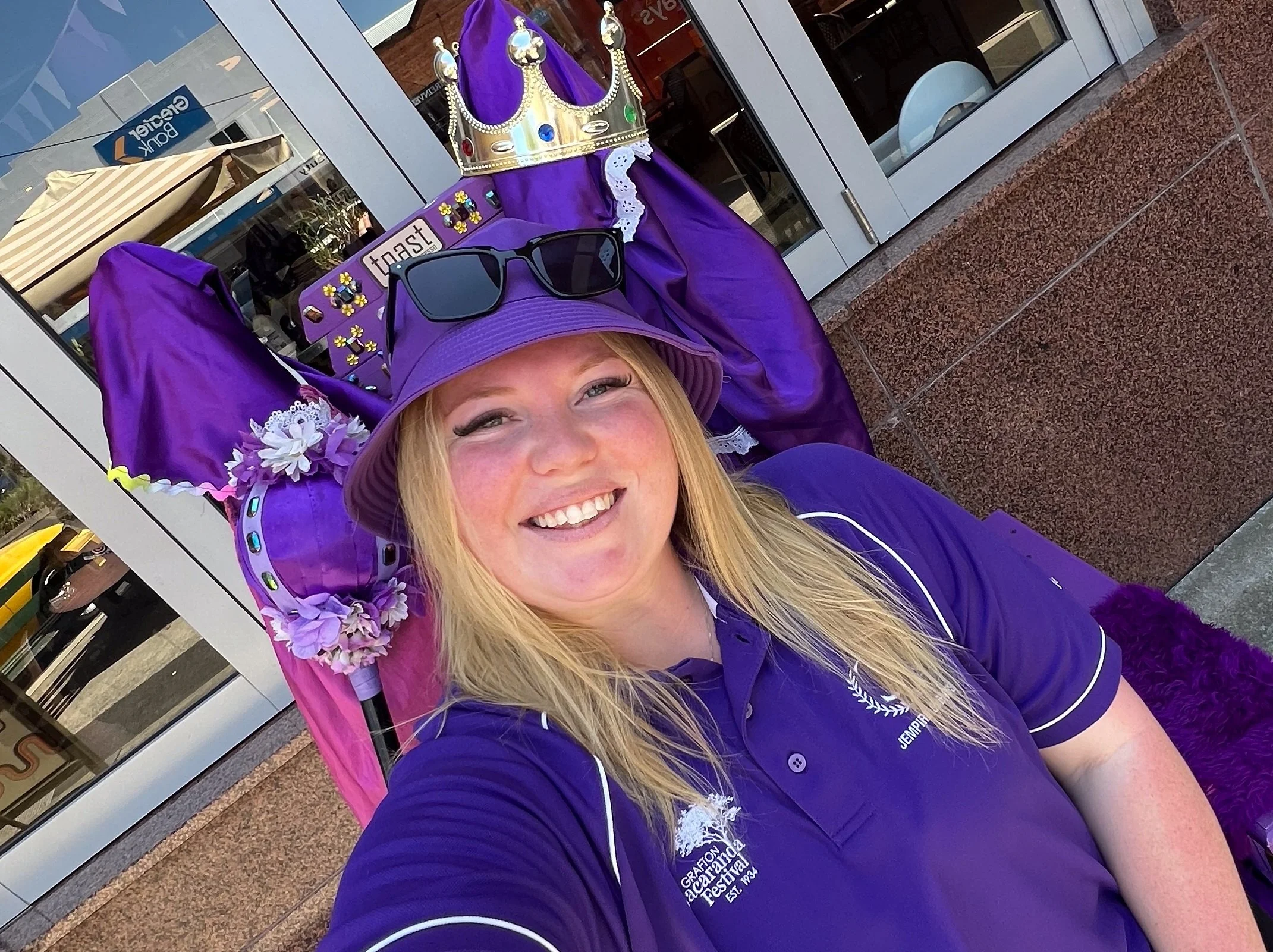 A smiling woman with long blonde hair wearing sunglasses, a purple hat, and a purple shirt with white lettering. She is sitting outside in front of a building with glass doors, and there's a festive display behind her that includes a purple cape with a crown on top and various decorative accessories.