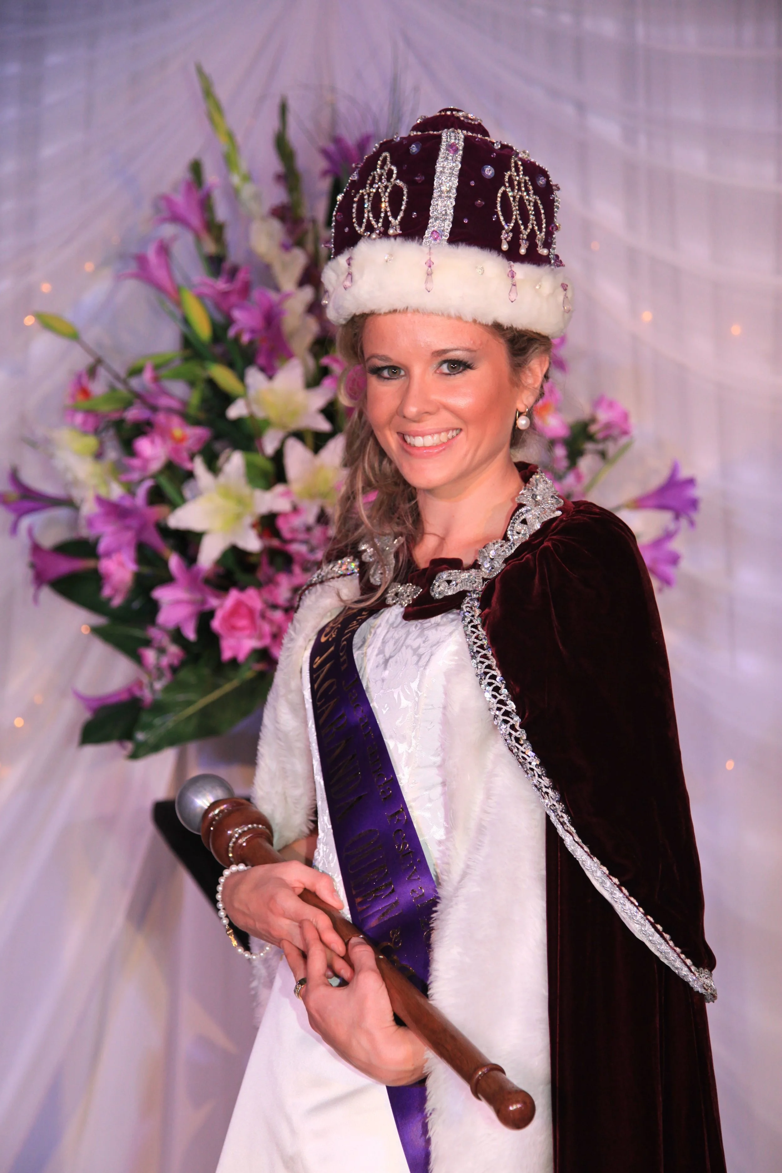 A woman wearing a crown, velvet robe, and sash, smiling at a celebration or pageant, with a bouquet of pink and white flowers in the background, and holding an over-sized gavel.