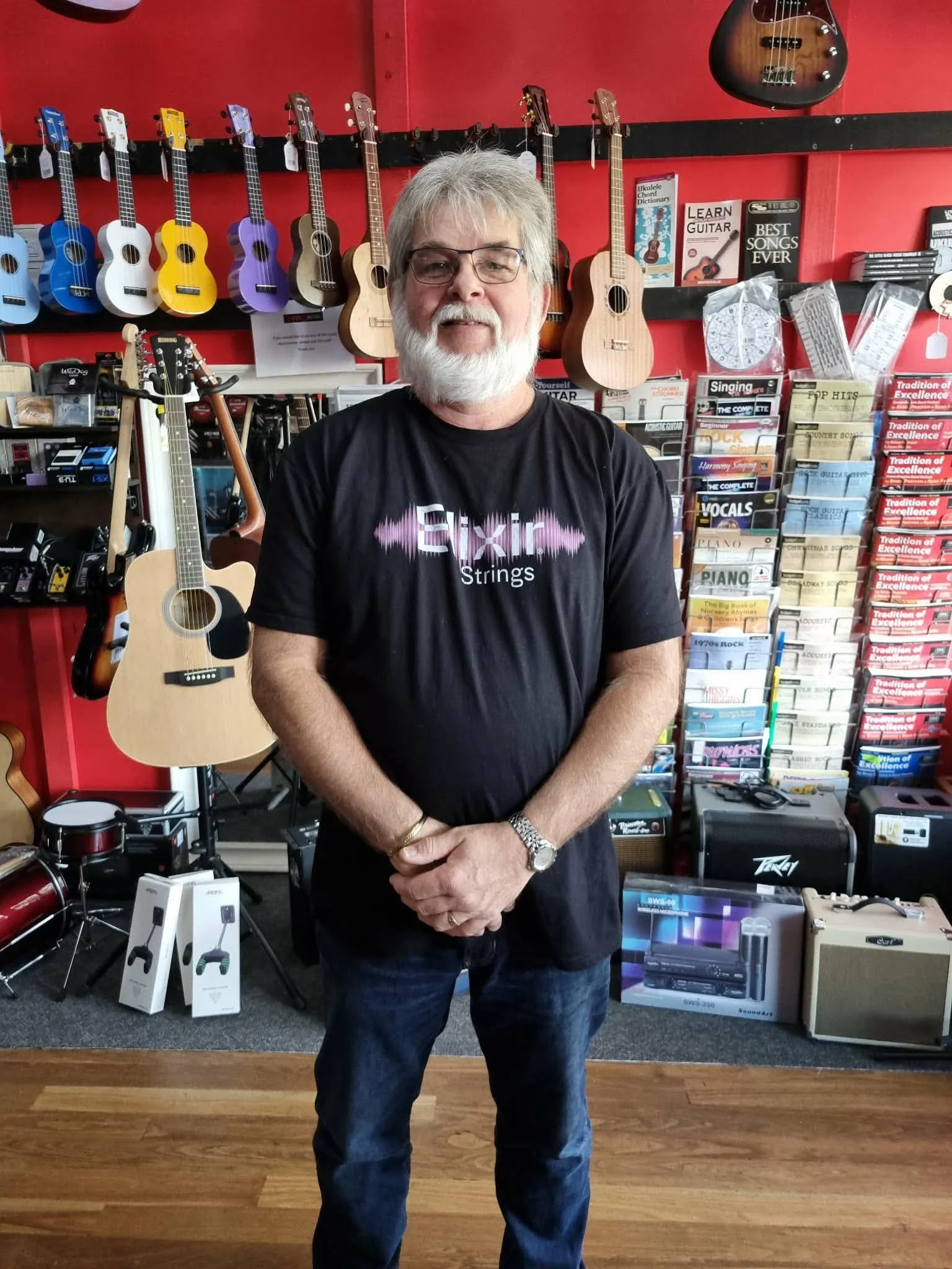 A man with gray hair, glasses, and a beard wearing a black T-shirt with a pink and white logo stands inside a music store. The store has red walls, with ukuleles hanging on the wall behind him in various colors, and music books and audio equipment displayed on shelves and tables.