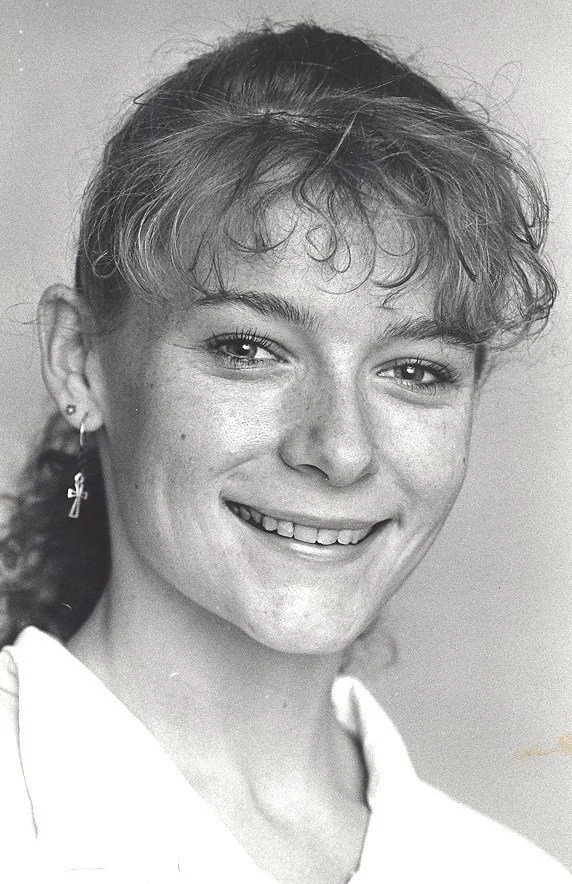 Black and white portrait of a young woman with curly hair, smiling, wearing earrings, and a white top.