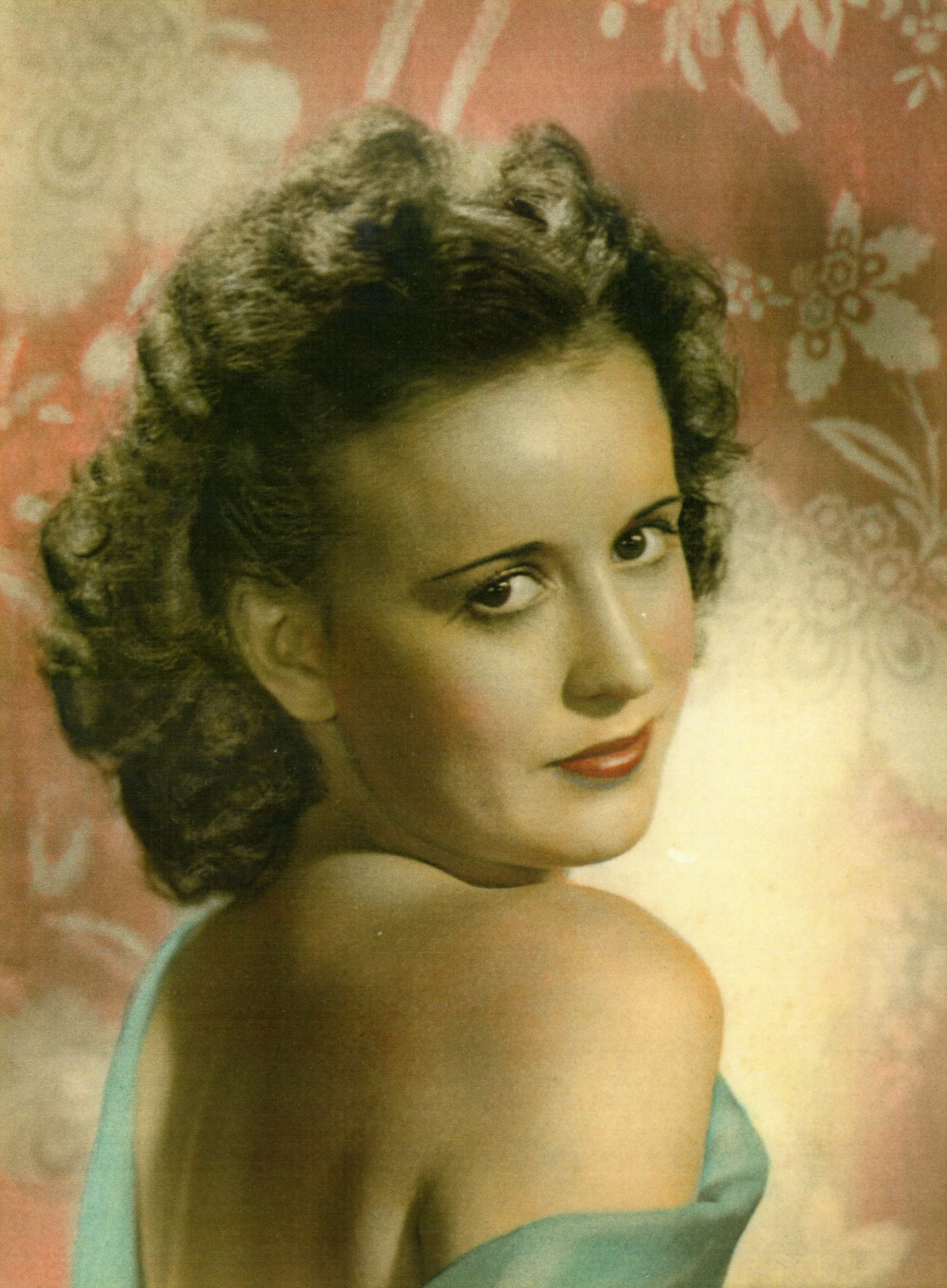 Close-up of a woman's face and shoulder, with styled curly hair, wearing red lipstick and a turquoise top, against a pink and floral background.
