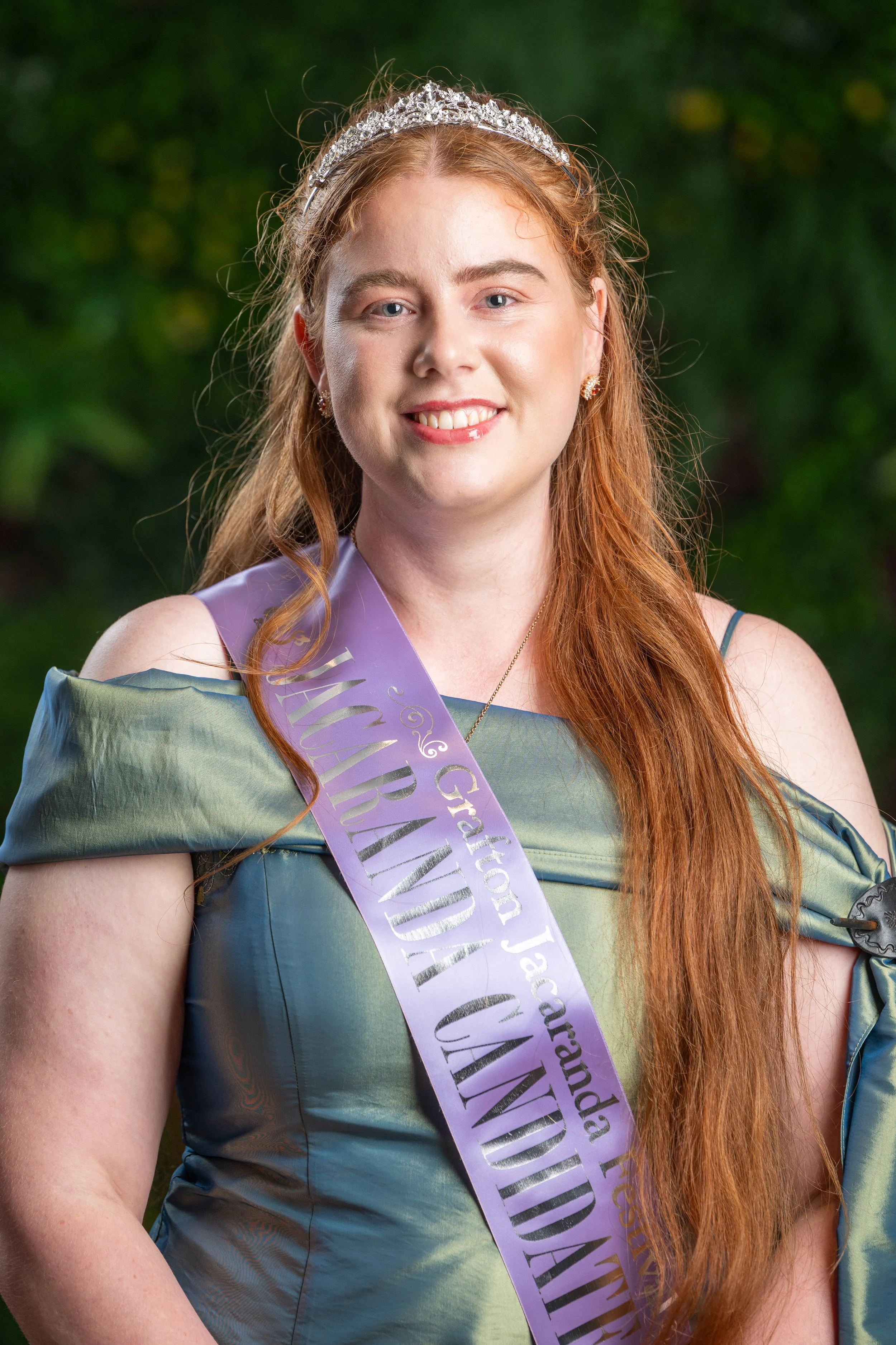 Young woman with red hair wearing a crown and a purple sash, smiling, outdoors with a leafy background.