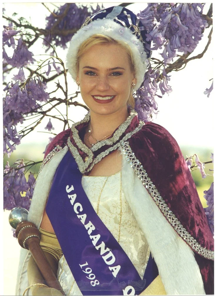 Young woman dressed in royal attire, wearing a crown and sash that reads 'JACKARANDA 1998', standing in front of purple flowering bush.