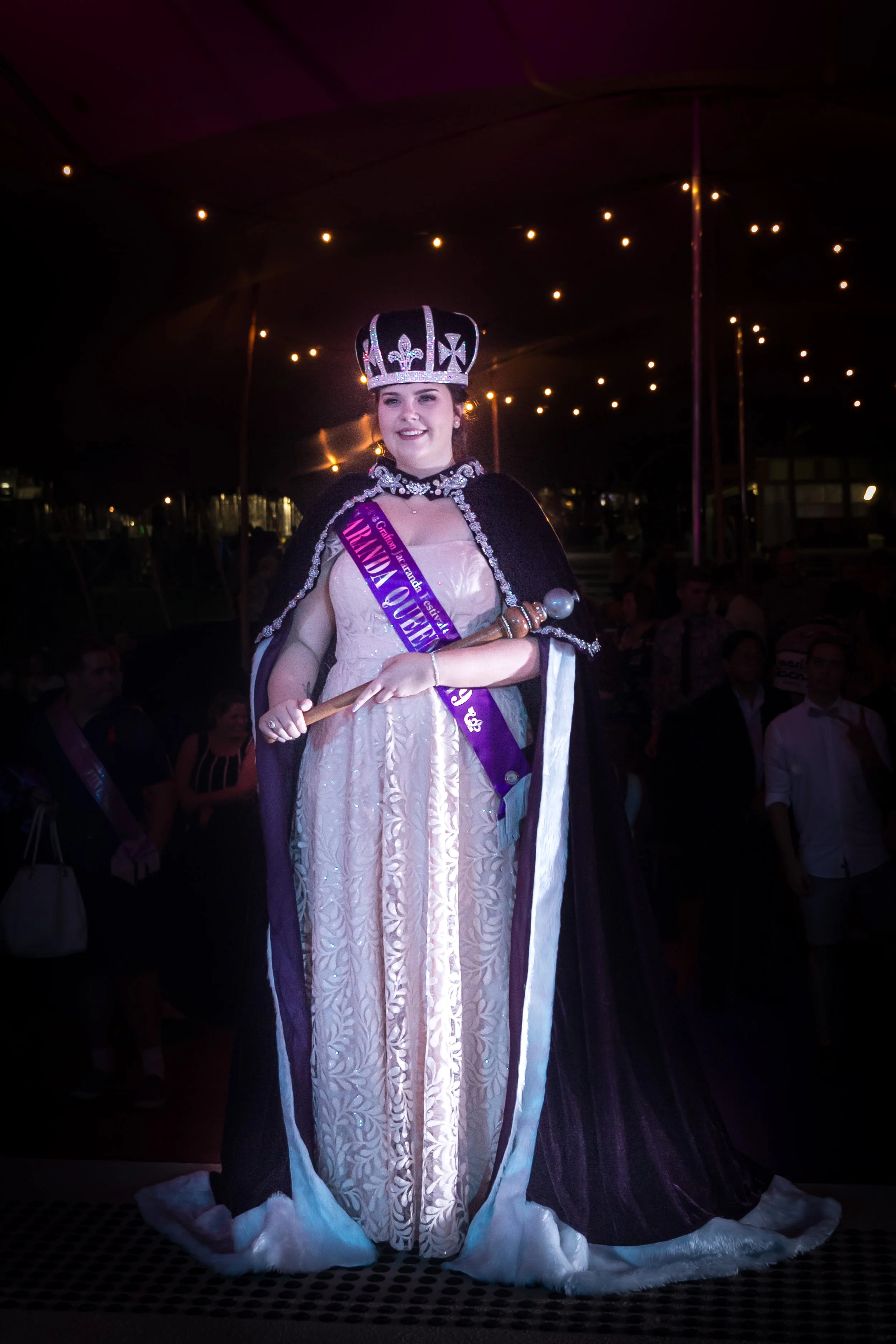Young woman dressed as royalty with a tiara, crown, and sash reading "TRENDA QUEEN" standing in front of a dark background with string lights.