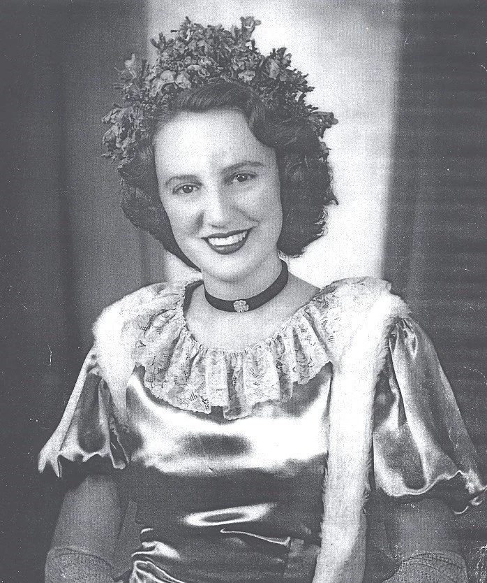 A black and white portrait of a woman with curly hair wearing a floral crown, a satin dress with ruffled collar, and a choker necklace.