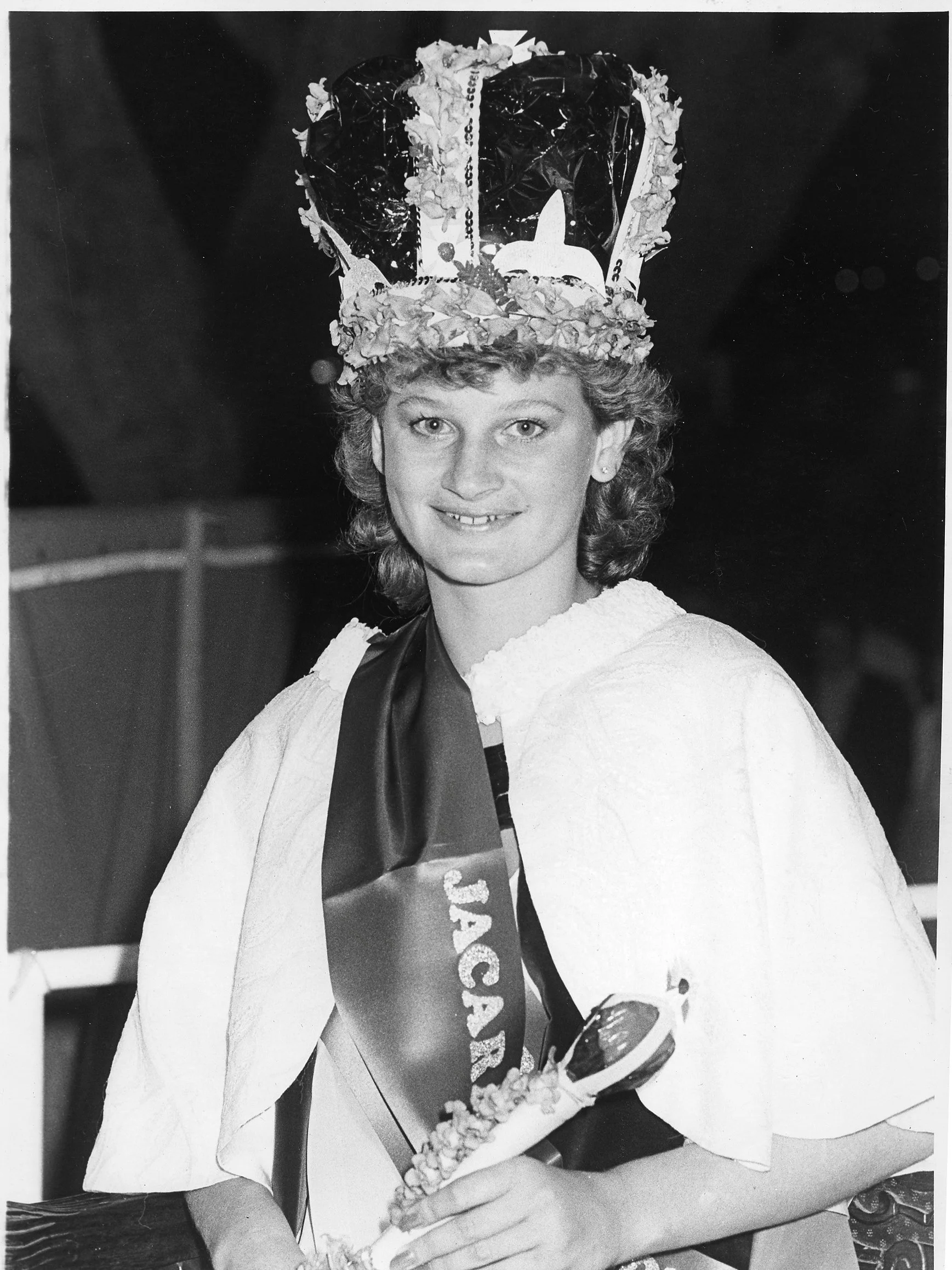 Black and white photo of a young woman wearing a school graduation sash and a decorated cap, smiling at the camera.
