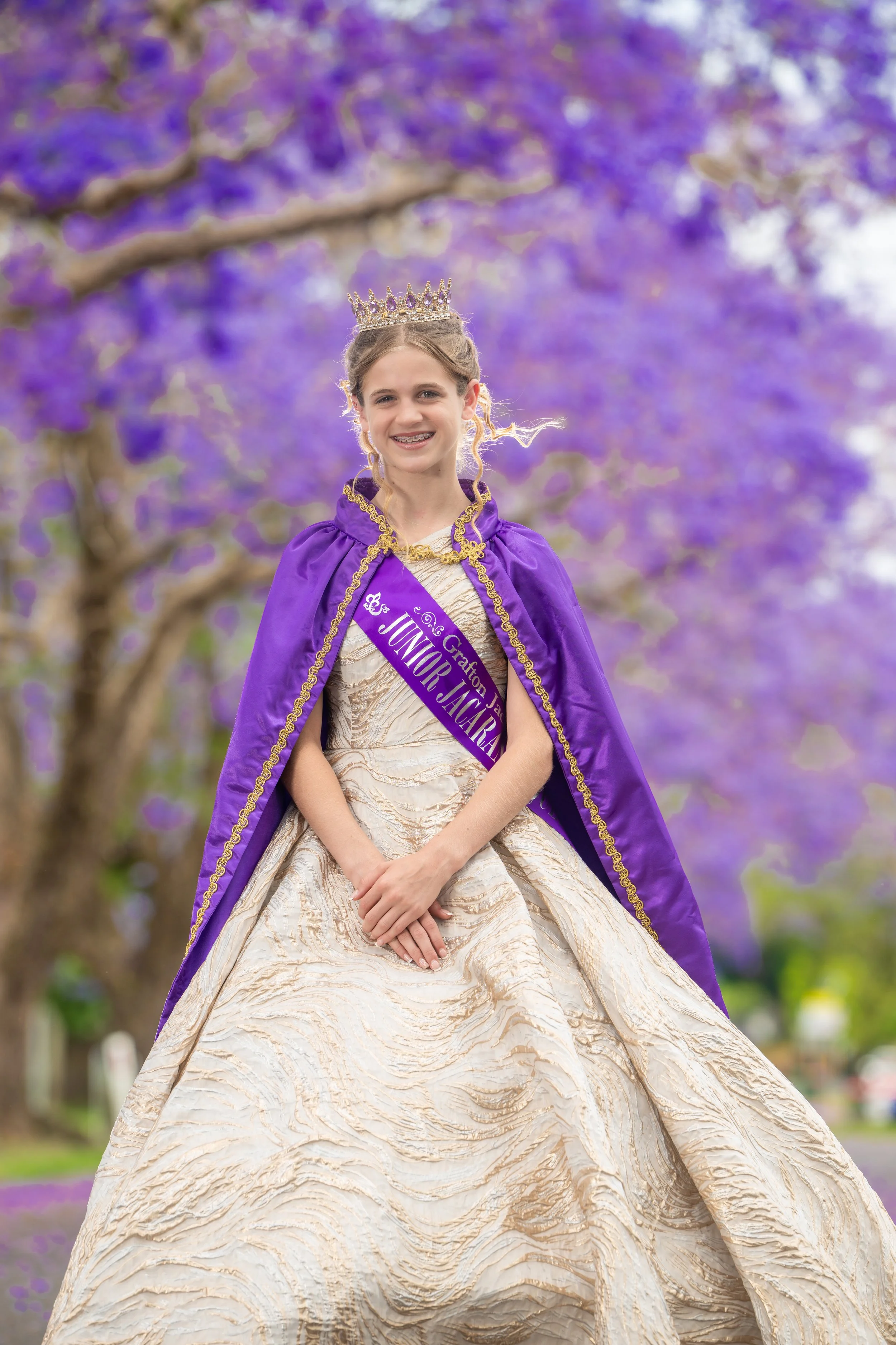 A young girl dressed as a queen or princess, wearing a gold and cream gown, a purple sash that says 'Junior' and a purple cape with gold trim, and a tiara, standing outdoors in front of purple flowering trees.