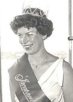 Black and white photo of a woman wearing a sash that reads 'Queen' and a crown, smiling.