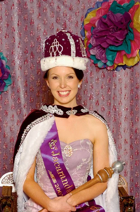 A woman wearing a crown, sash, and cape, sitting on a throne, smiling, with colorful paper decorations in the background.
