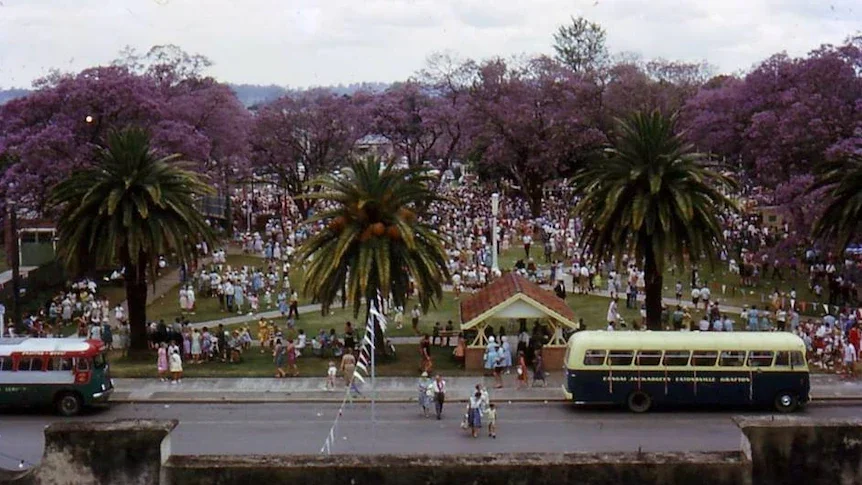 Park filled with people, many in white, under pink-flowering trees, with palm trees, a small building, and a bus on the street in front.
