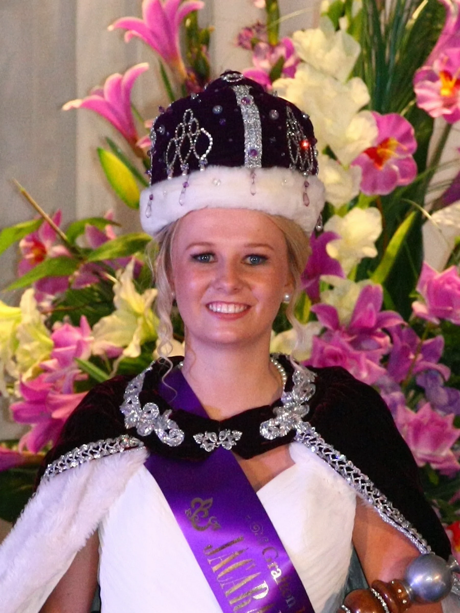 Woman wearing a crown and a sash, celebrating a victory, surrounded by pink and white flowers.