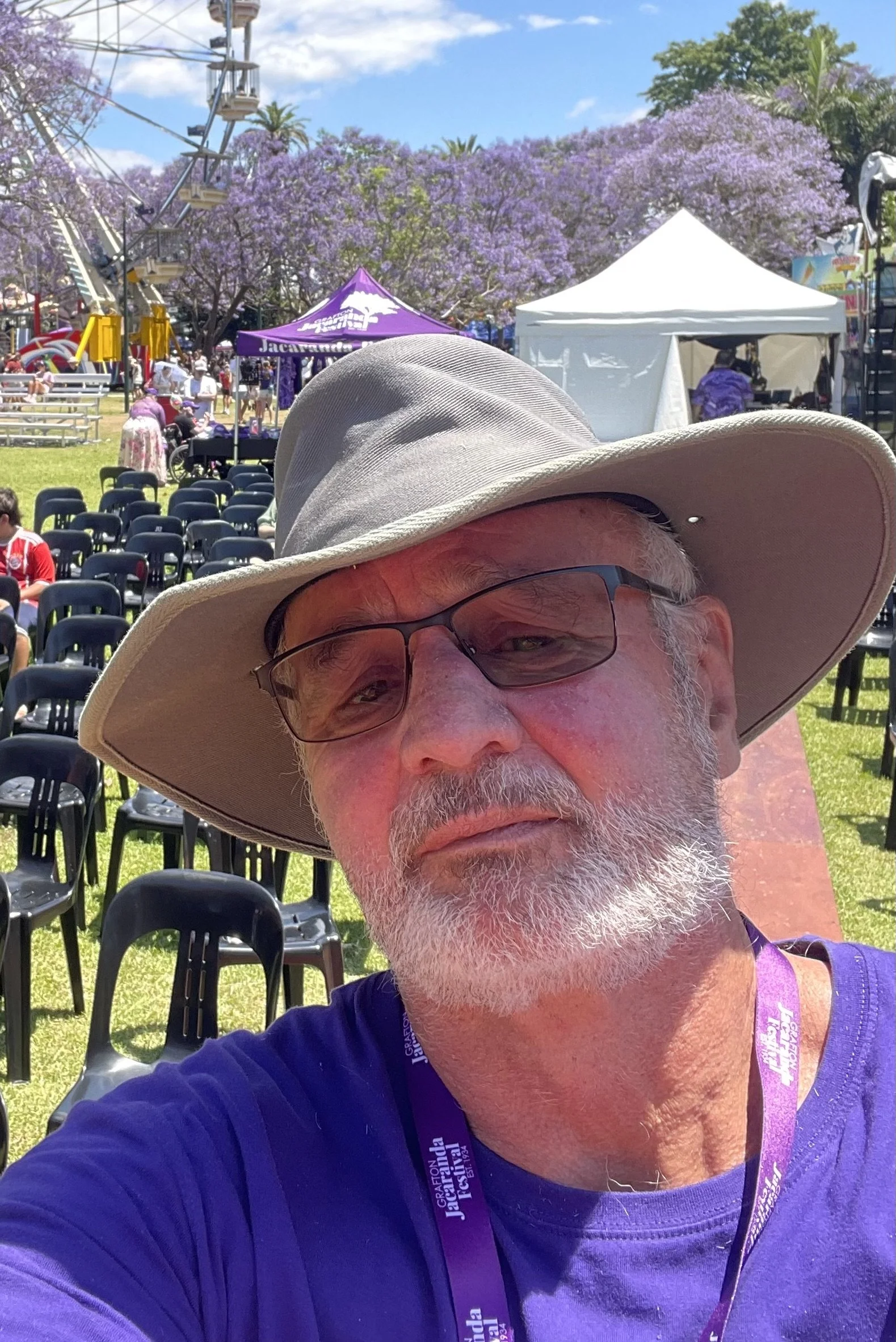 A man with glasses and a wide-brimmed hat taking a selfie at an outdoor event with purple trees, carnival rides, tents, and chairs in the background.