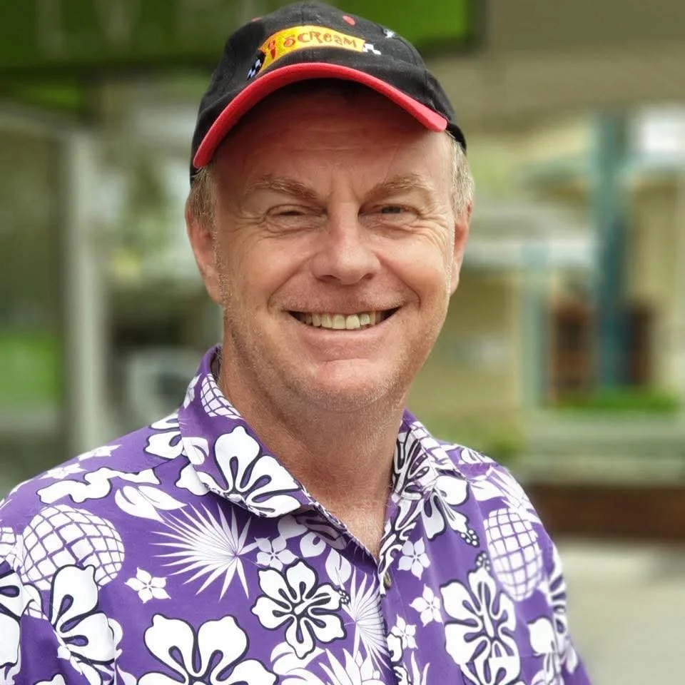 A smiling older man wearing a black baseball cap with a red brim, a purple Hawaiian shirt with white floral pattern, and standing outdoors with a blurred background of green and brown scenery.