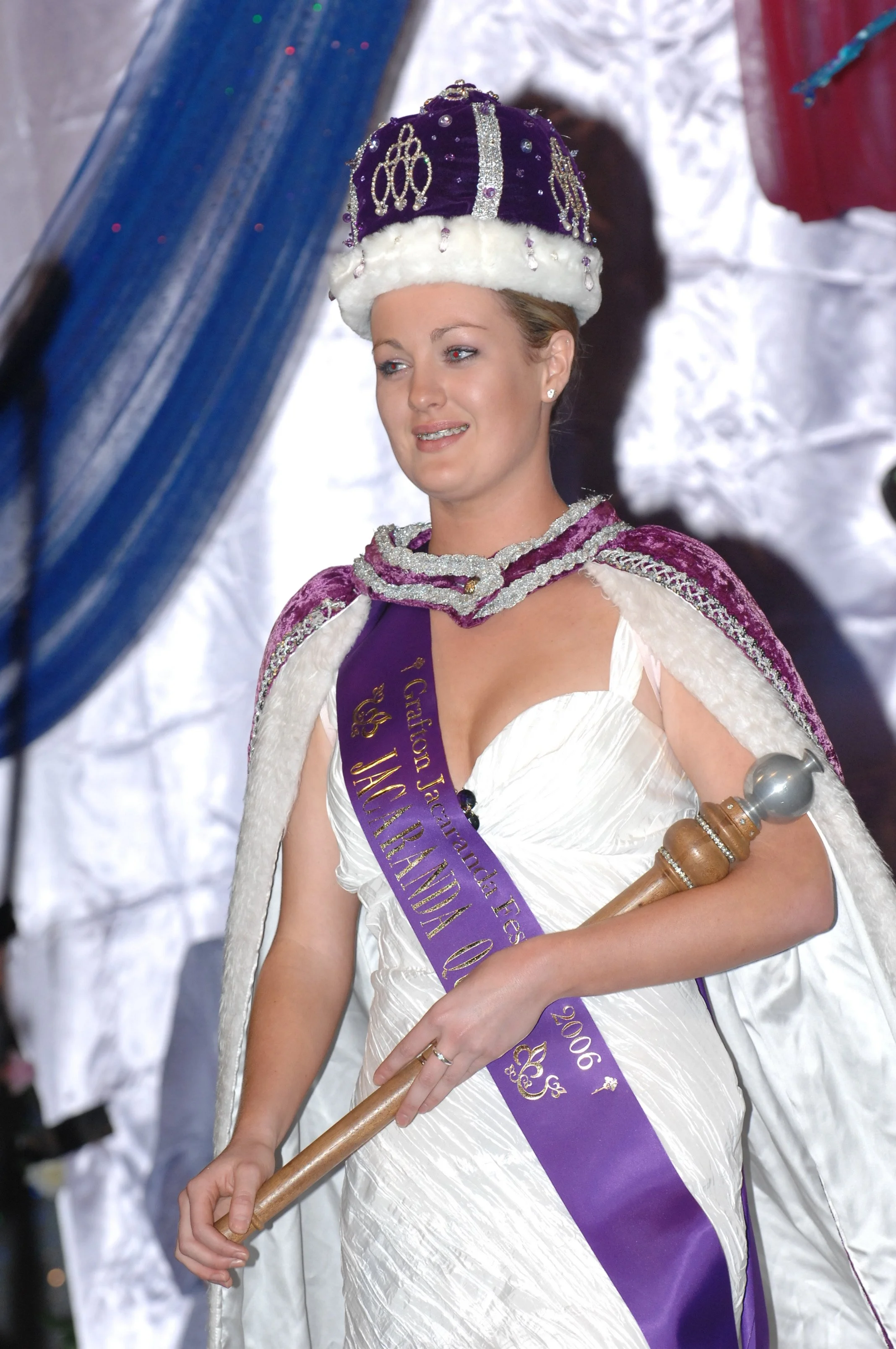 Woman wearing a king or queen crown with purple velvet, white fur trim, and jeweled embellishments, dressed in a white gown and purple sash, holding a scepter, at a parade or celebration, likely a Mardi Gras or similar event.