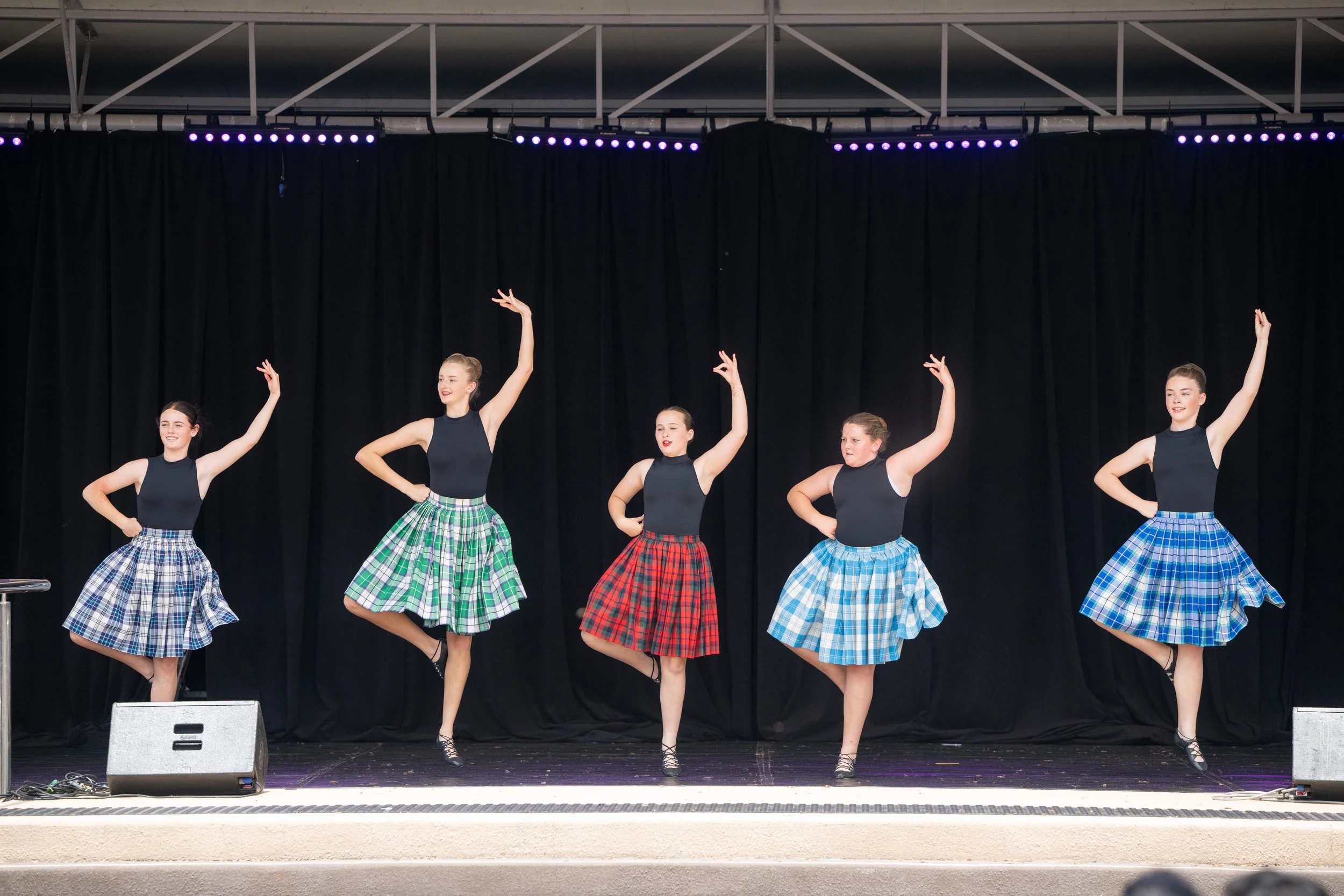 Group of five young women in plaid skirts and black tops performing a dance on stage with a black curtain backdrop and purple stage lights.