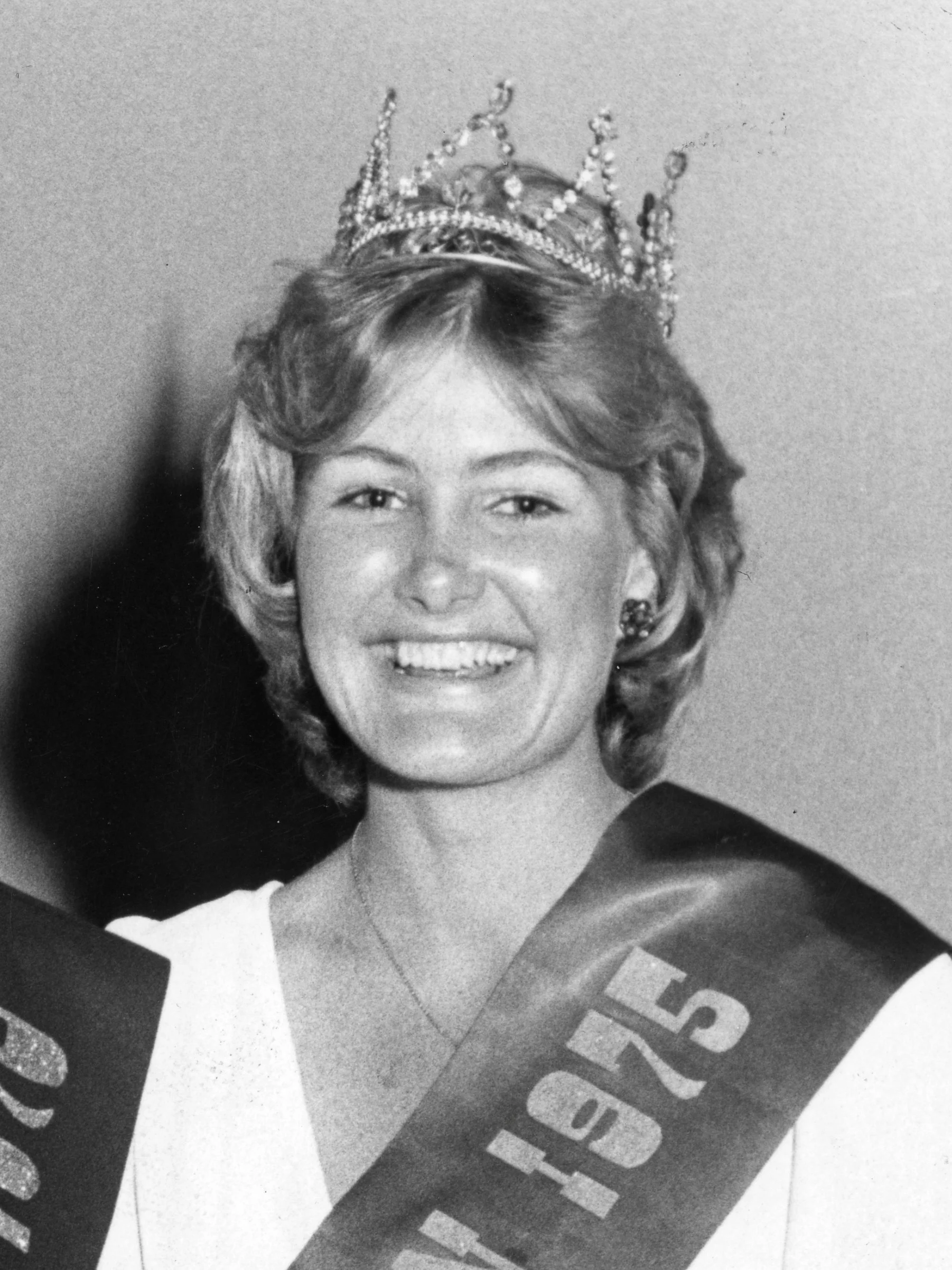 Black and white photo of a woman with short wavy hair wearing a crown or tiara and earrings, holding a piece of cloth with partially visible text.