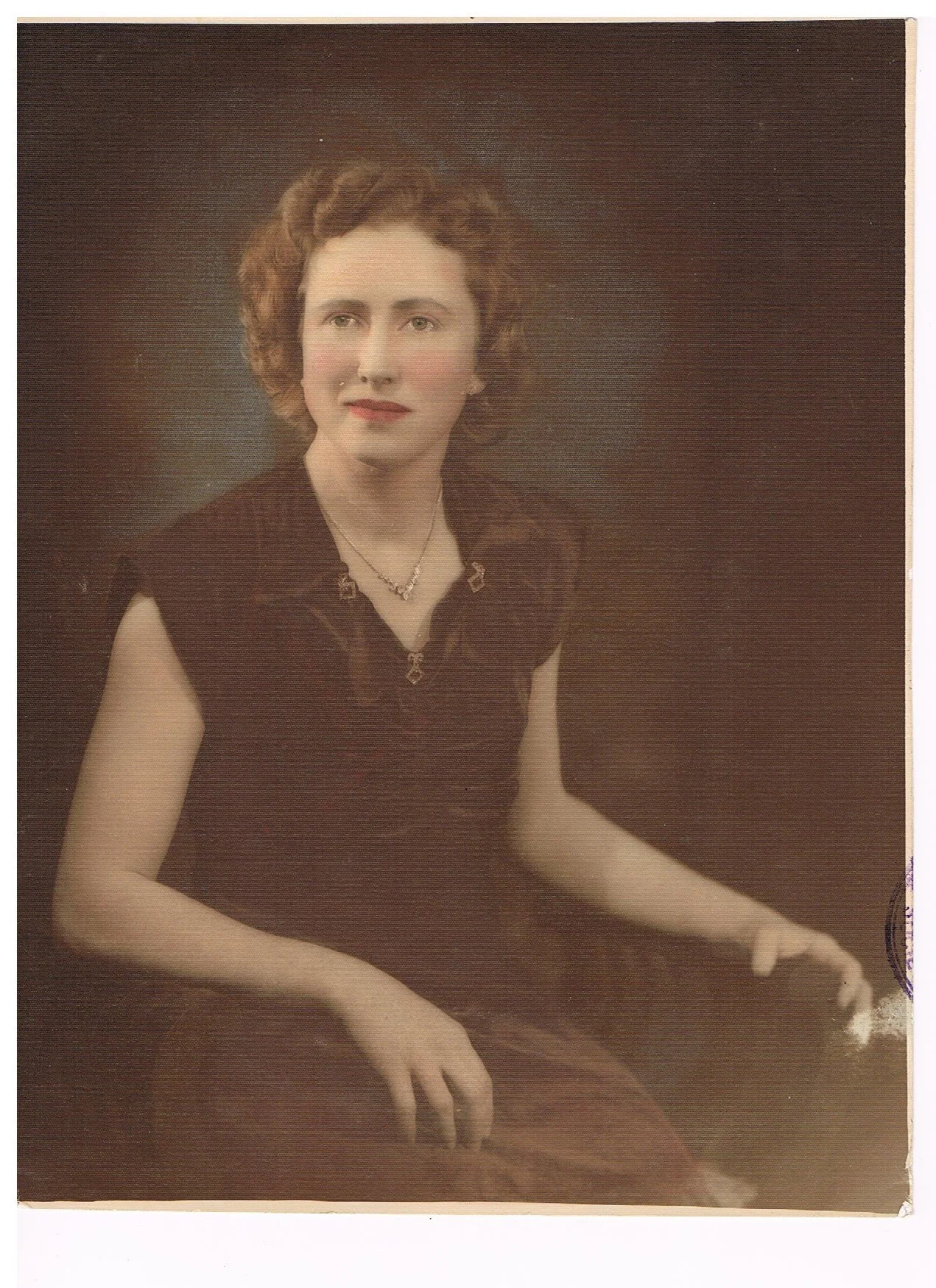 A woman with curly hair, wearing a dark top with short sleeves, jewelry, and sitting against a dark background.