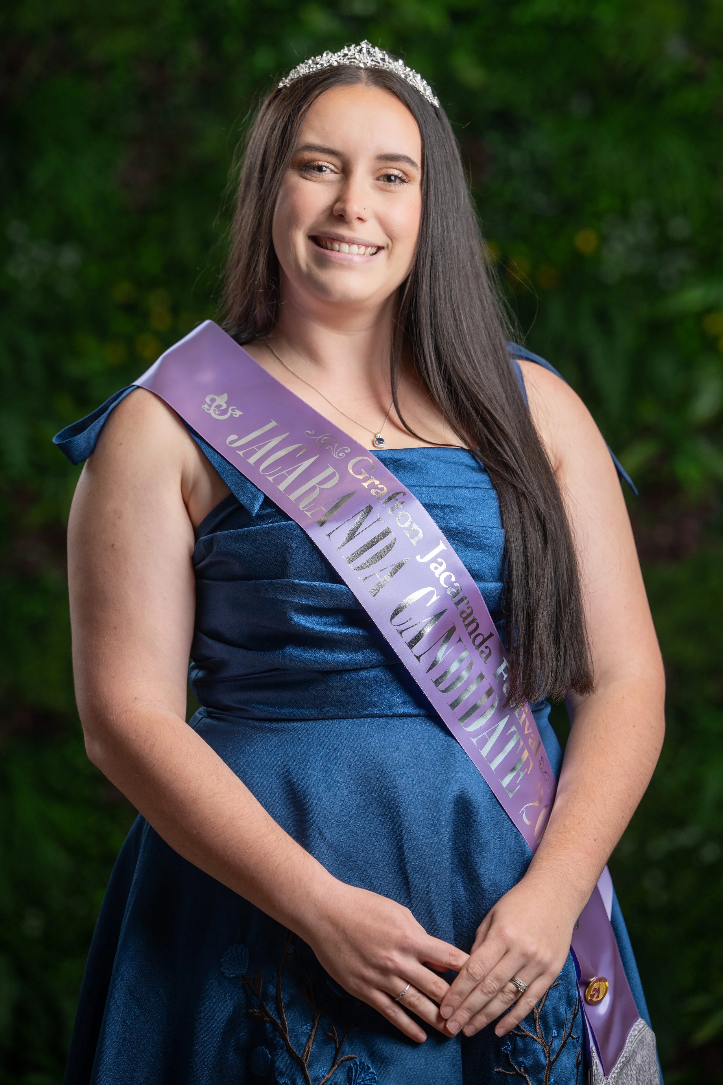 A young woman with long dark hair, wearing a tiara and a blue dress, smiling, standing in front of a greenery background, with a lavender sash that says 'Graduation Jack...'