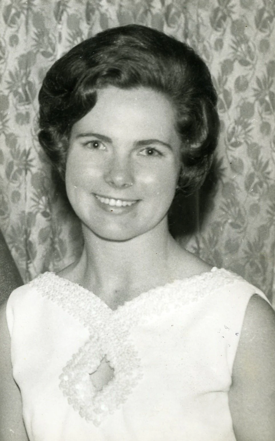 Black and white portrait of a woman with styled hair, smiling, wearing a sleeveless dress with lace details, standing in front of a patterned curtain.