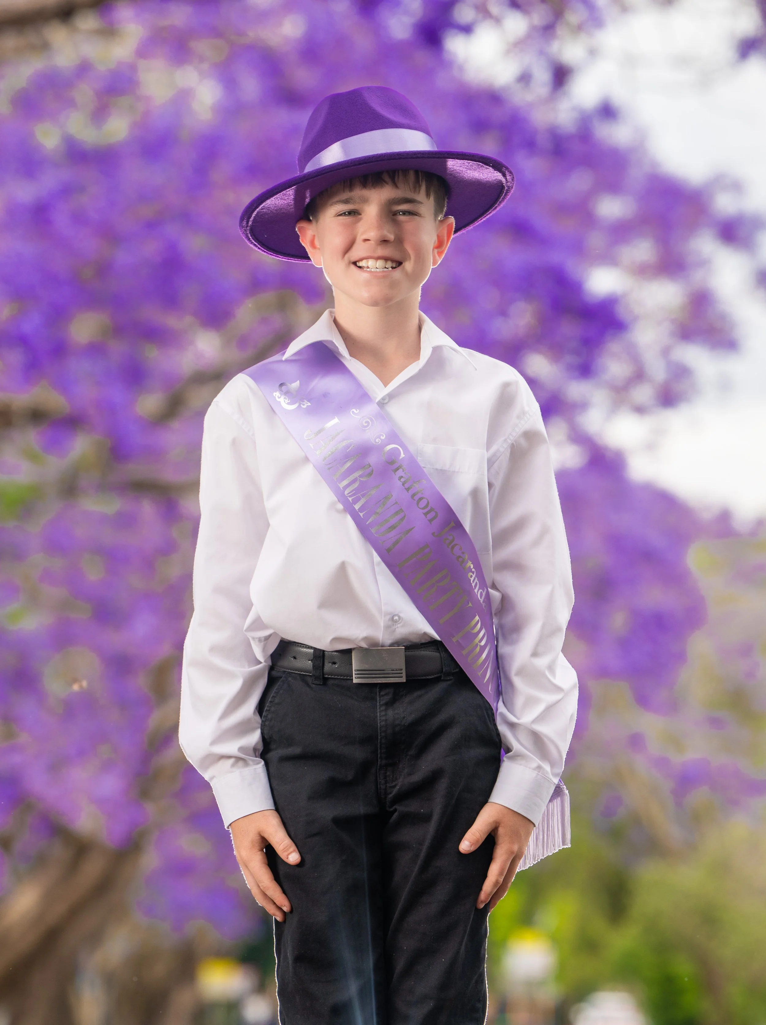A young man wearing a purple hat and a sash, standing outdoors with purple flowering trees in the background.