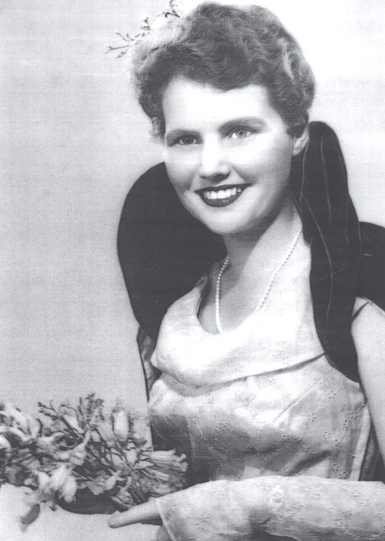 Black and white portrait of a woman with short curly hair, smiling, wearing a pearl necklace, holding a bouquet of flowers, dressed in elegant clothing, with a dark bow or ribbon on her shoulder, and a light-colored dress with lace or embroidery.