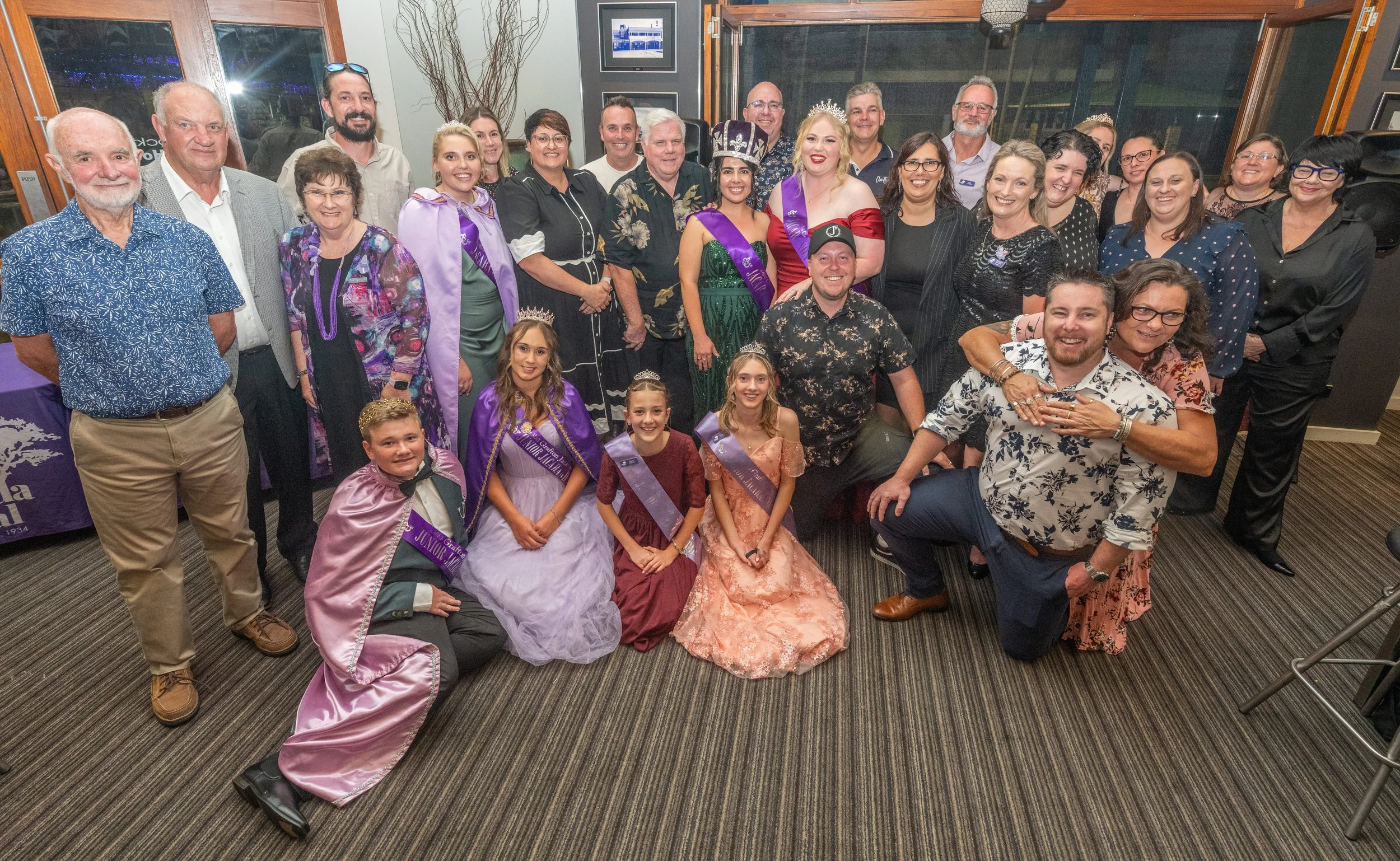 A large group of adults and children at a celebration or pageant, with some children wearing sashes and crowns, and a few women wearing tiaras and sashes, inside a decorated indoor venue.
