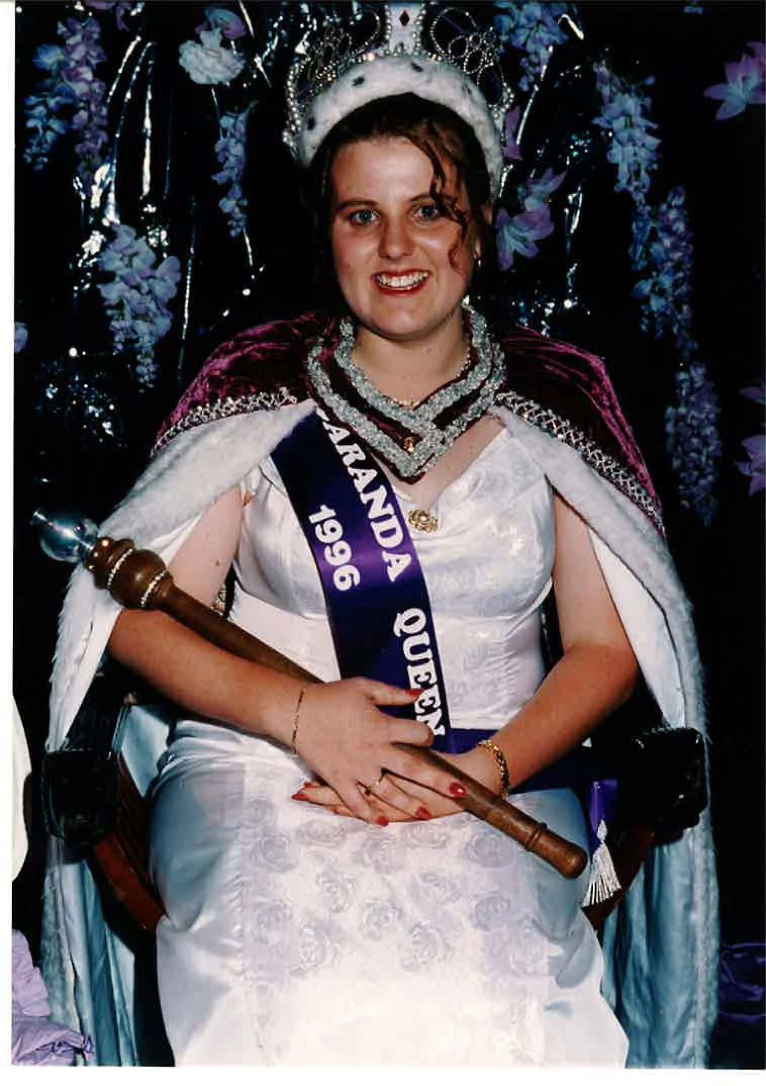 A woman dressed as a queen, sitting on a throne with a crown and a sash that reads "Queen 96." She has curly brown hair, blue eyes, and is wearing a white lace dress with jewelry, including necklaces, bracelets, and rings. She is holding a wooden scepter, and the background has purple and blue floral decorations.