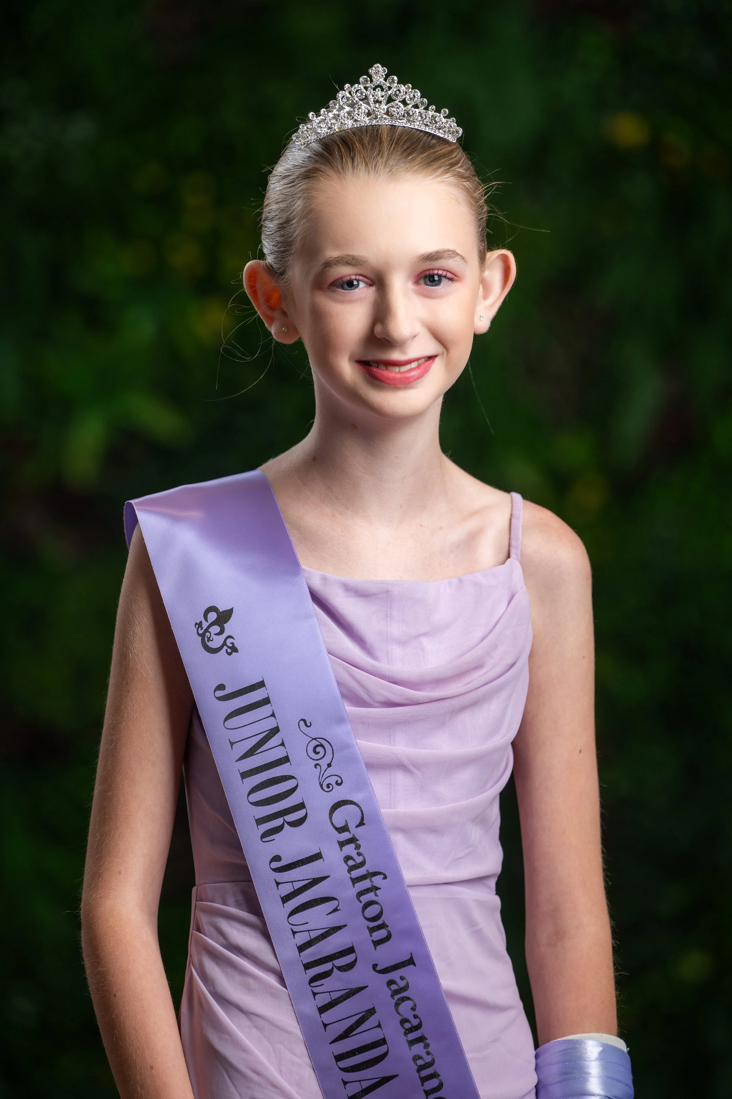 Young girl wearing a lavender dress and a tiara, smiling, with a lavender sash across her chest that reads 'JUNIOR J' and more text, standing outdoors with a blurred green background.