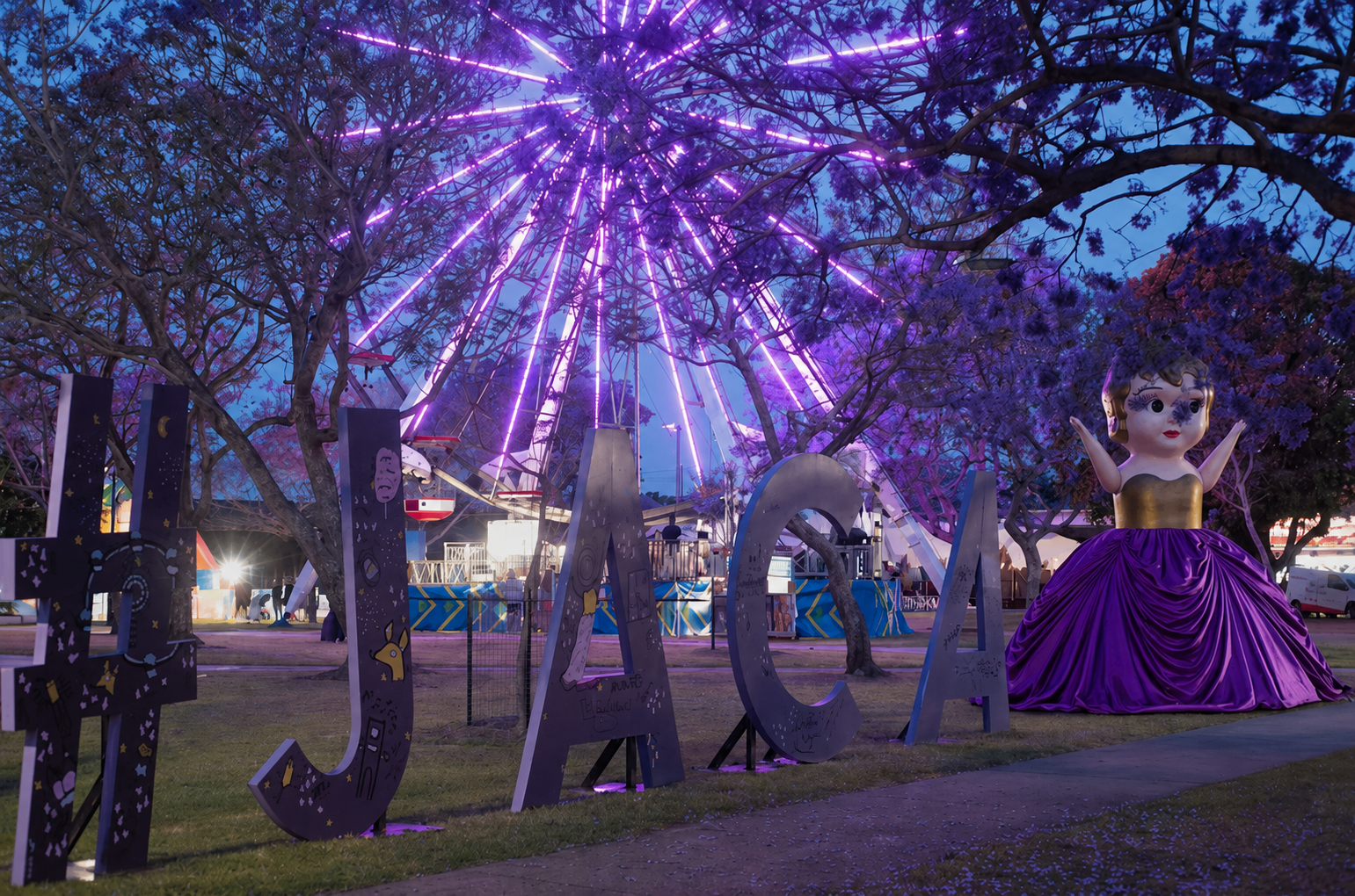 A nighttime scene at a carnival with a lit-up Ferris wheel in the background, surrounded by trees with purple blossoms. Large decorative letters spell out 'JUMA' in front of the trees. There's a large cartoonish statue of a girl in a purple dress with arms raised nearby.