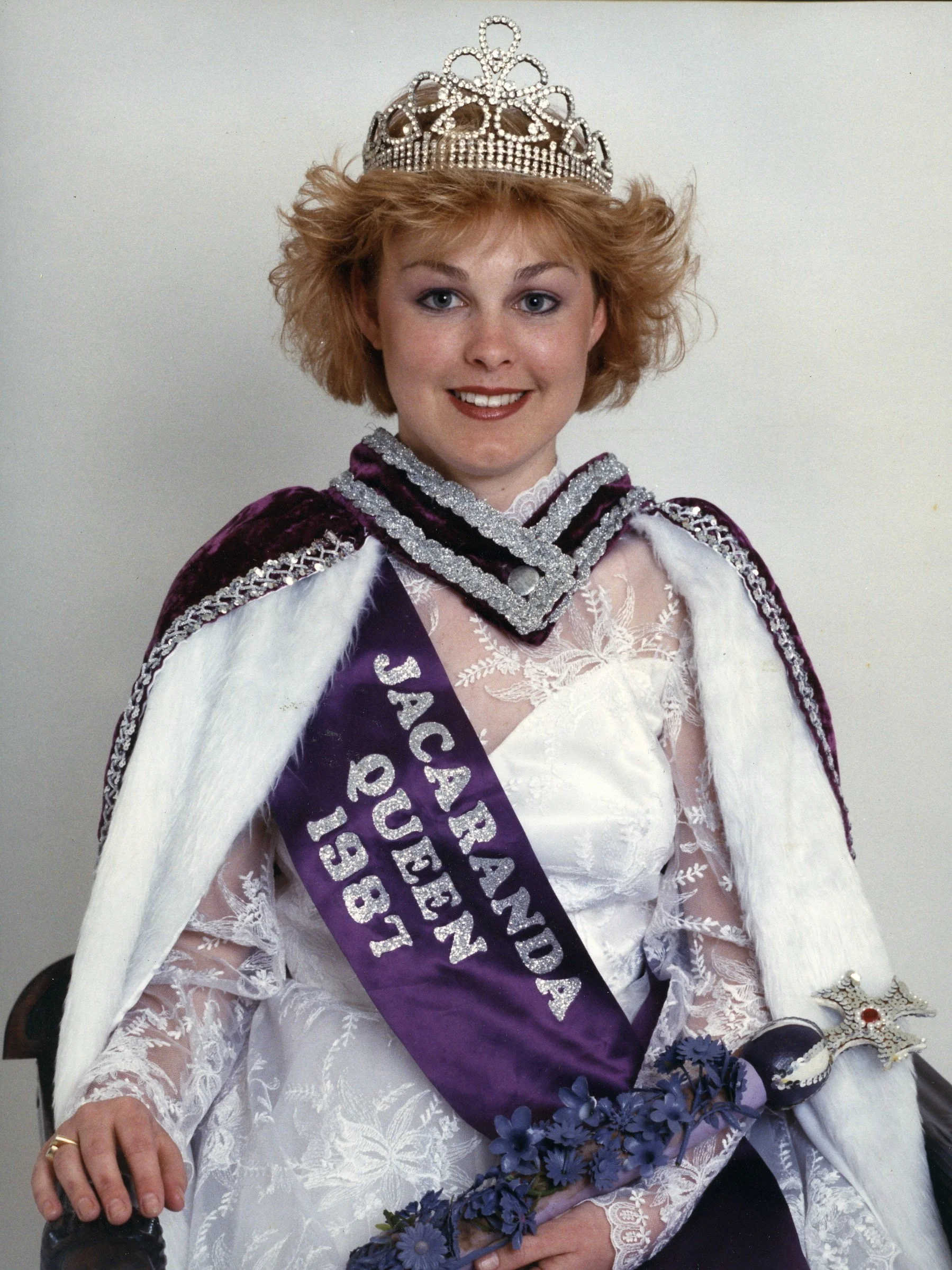 Woman wearing a tiara, sash that says 'Princess of the Year', and a white dress with lace details holding a bouquet of purple flowers.
