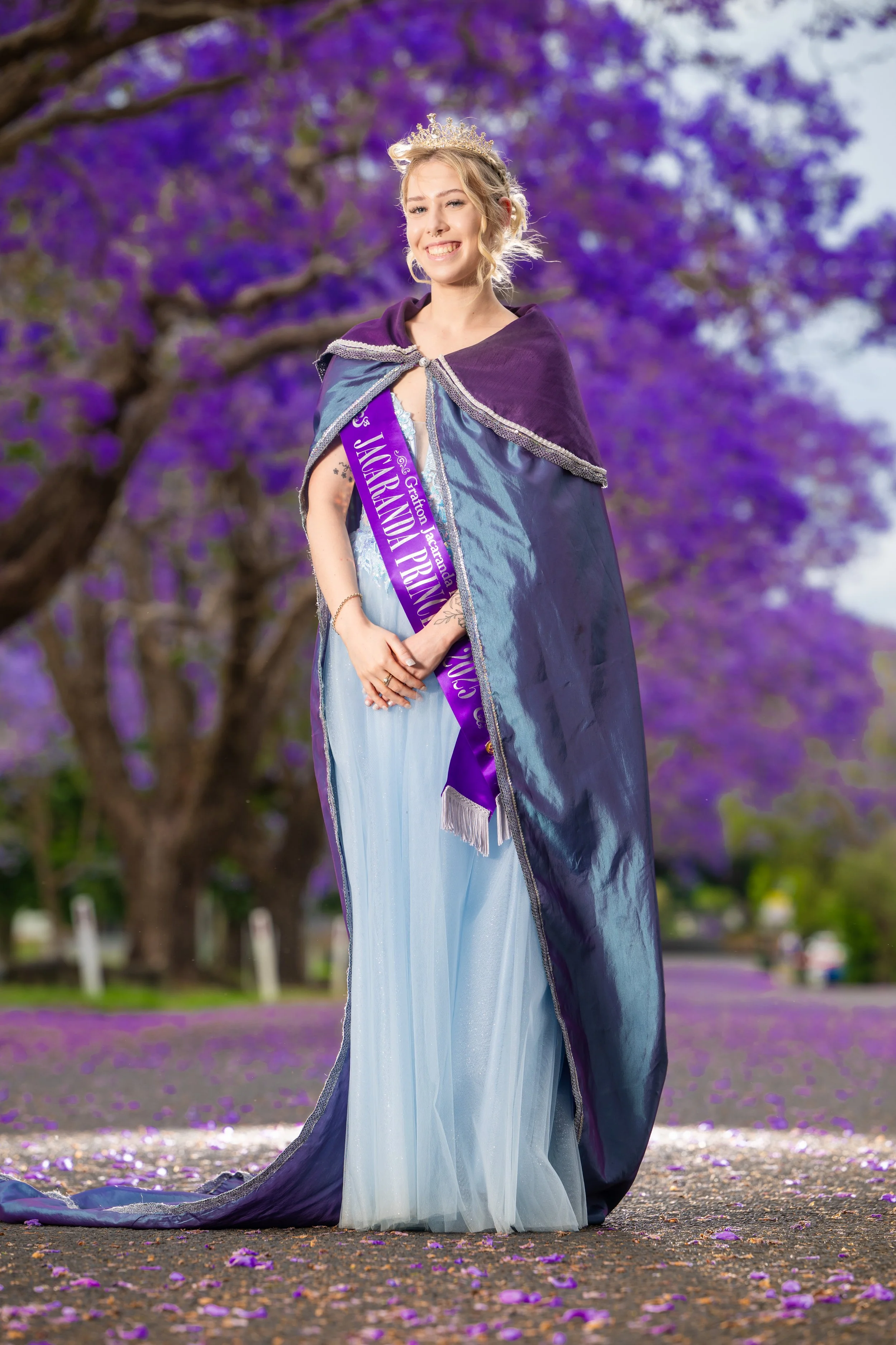 A woman wearing a crown and sash with the words 'JACARANDA PRINCESS' stands outdoors under a flowering jacaranda tree with purple blossoms, smiling and looking at the camera.