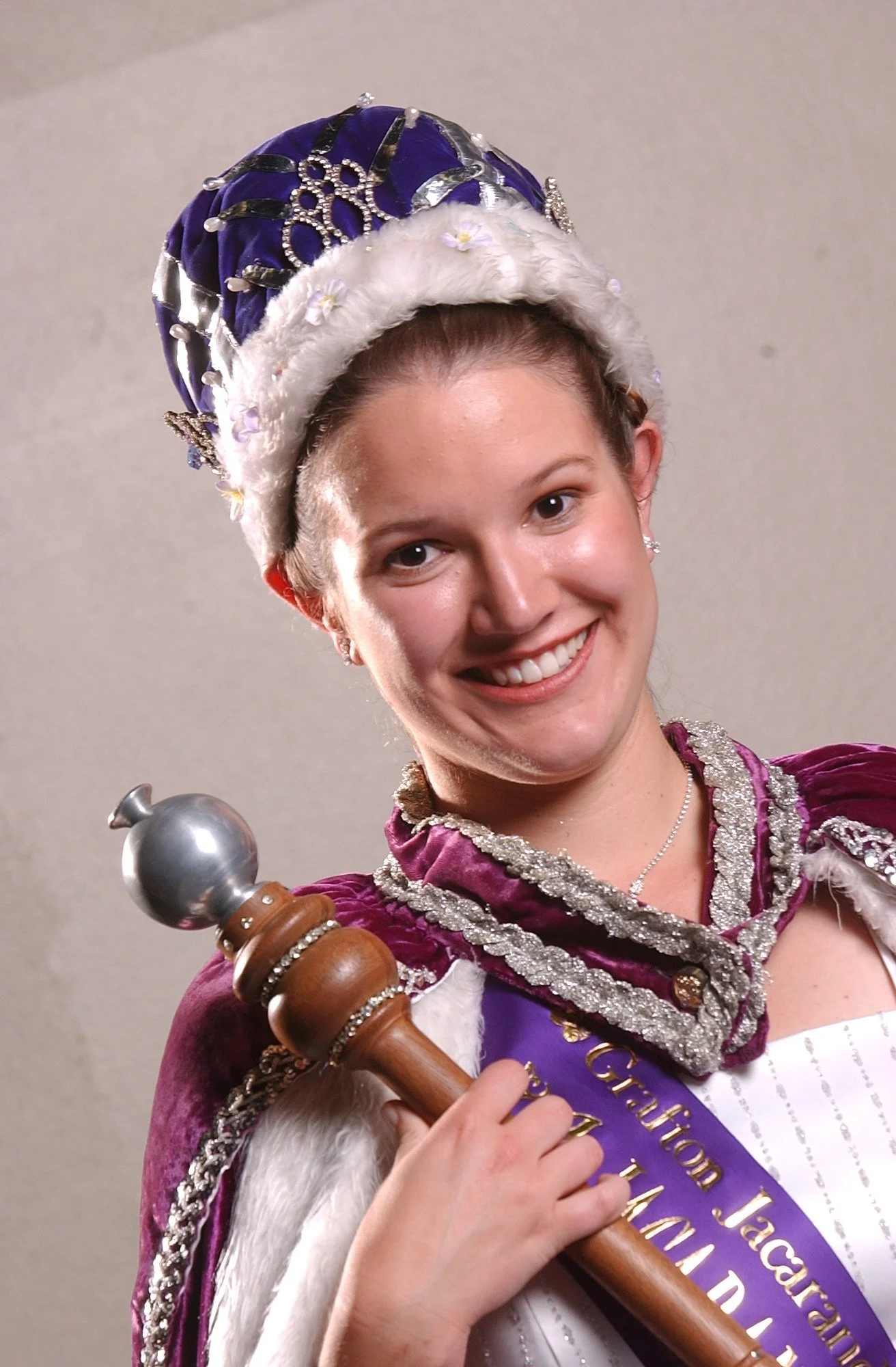 A woman dressed in royal costume, wearing a purple sash and a crown, holding a staff, smiling for the camera.