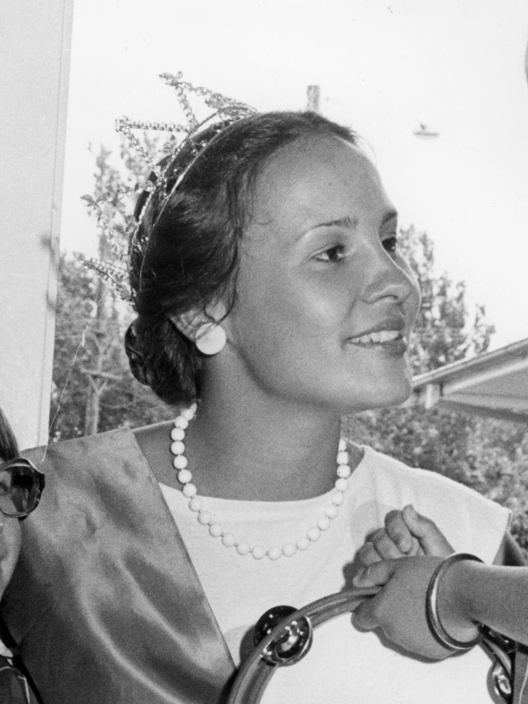 A black and white photo of a woman with short hair wearing a necklace, earrings, and a bracelet. She is smiling and touching her wrist with her hand. The background shows trees and what appears to be a tower or pole.
