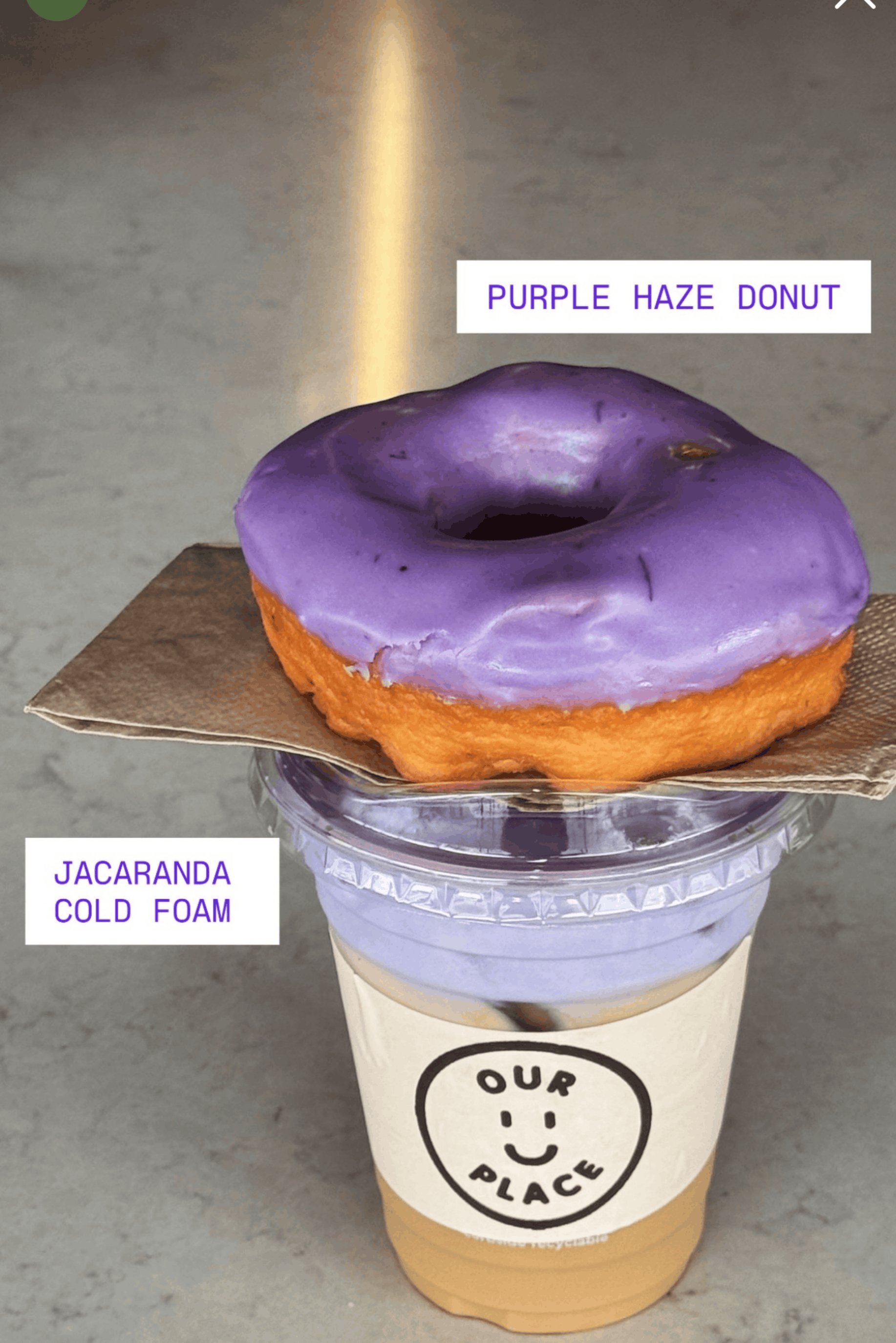 A donut with purple glaze labeled as 'Purple Haze Donut' sitting on top of a cup of jacaranda cold foam drink with the label 'Our Place' logo.
