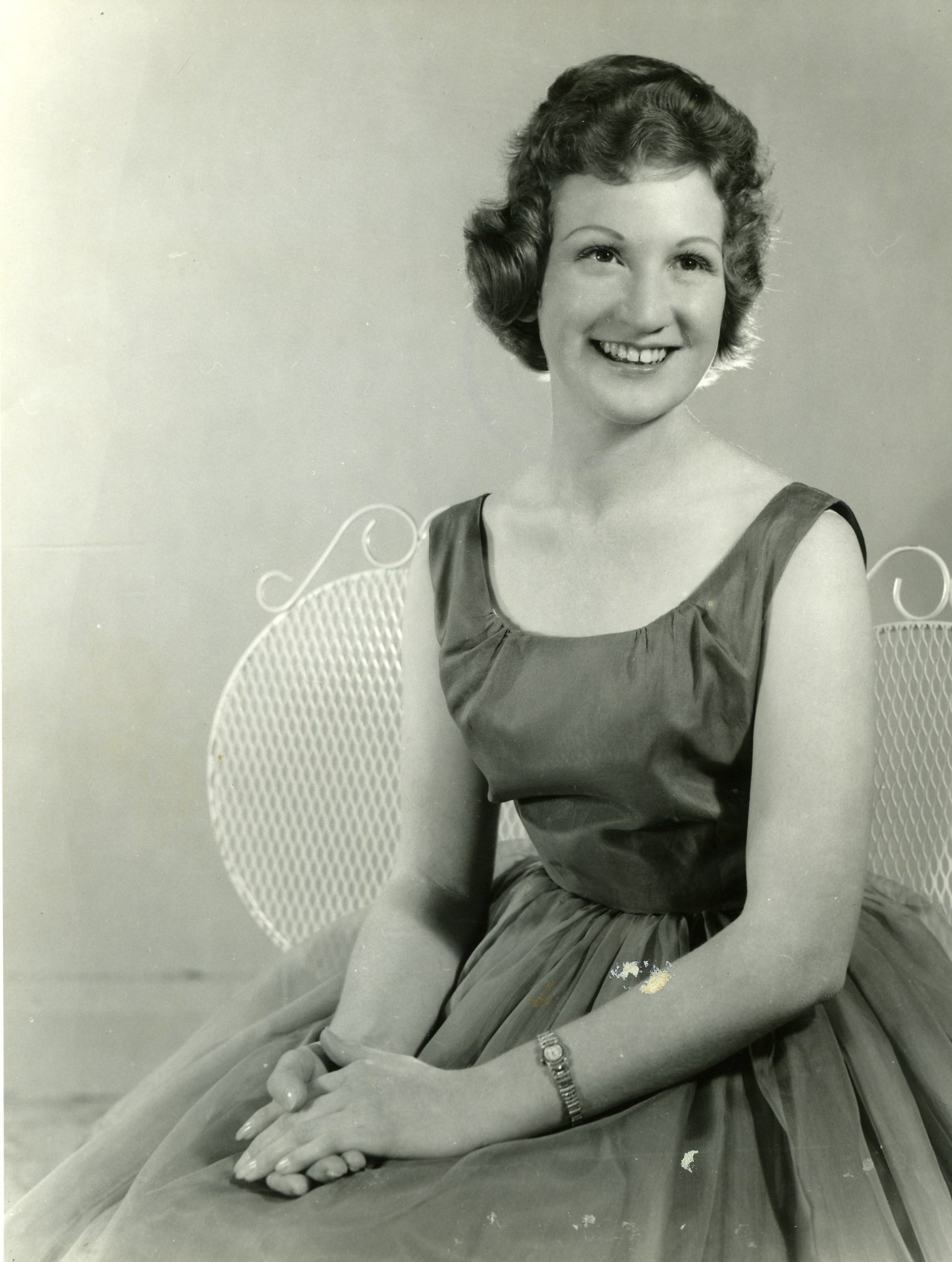 Black and white photo of a smiling woman with vintage hairstyle, wearing a sleeveless dress, sitting with her hands clasped on her lap.