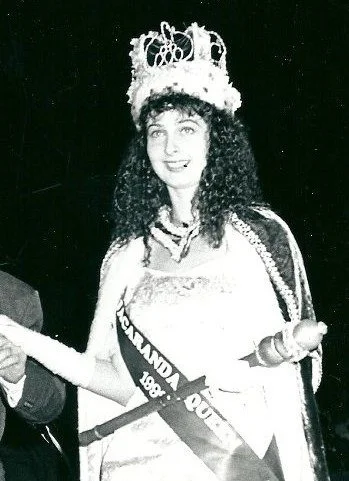 Black and white photo of a woman wearing a crown and sash, dressed as a beauty queen or pageant winner, holding a scepter.