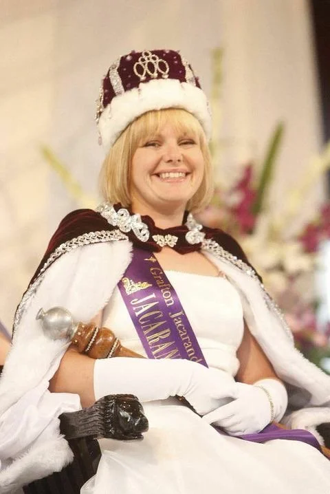 Woman smiling, dressed as a queen with a crown, royal sash, and cape, holding a scepter, in a setting with floral decorations.