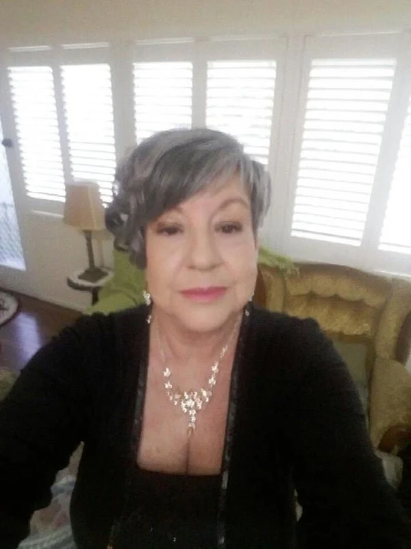 A woman with short gray hair taking a selfie in a living room with white plantation shutters on the windows, a green couch, and a gold upholstered armchair.