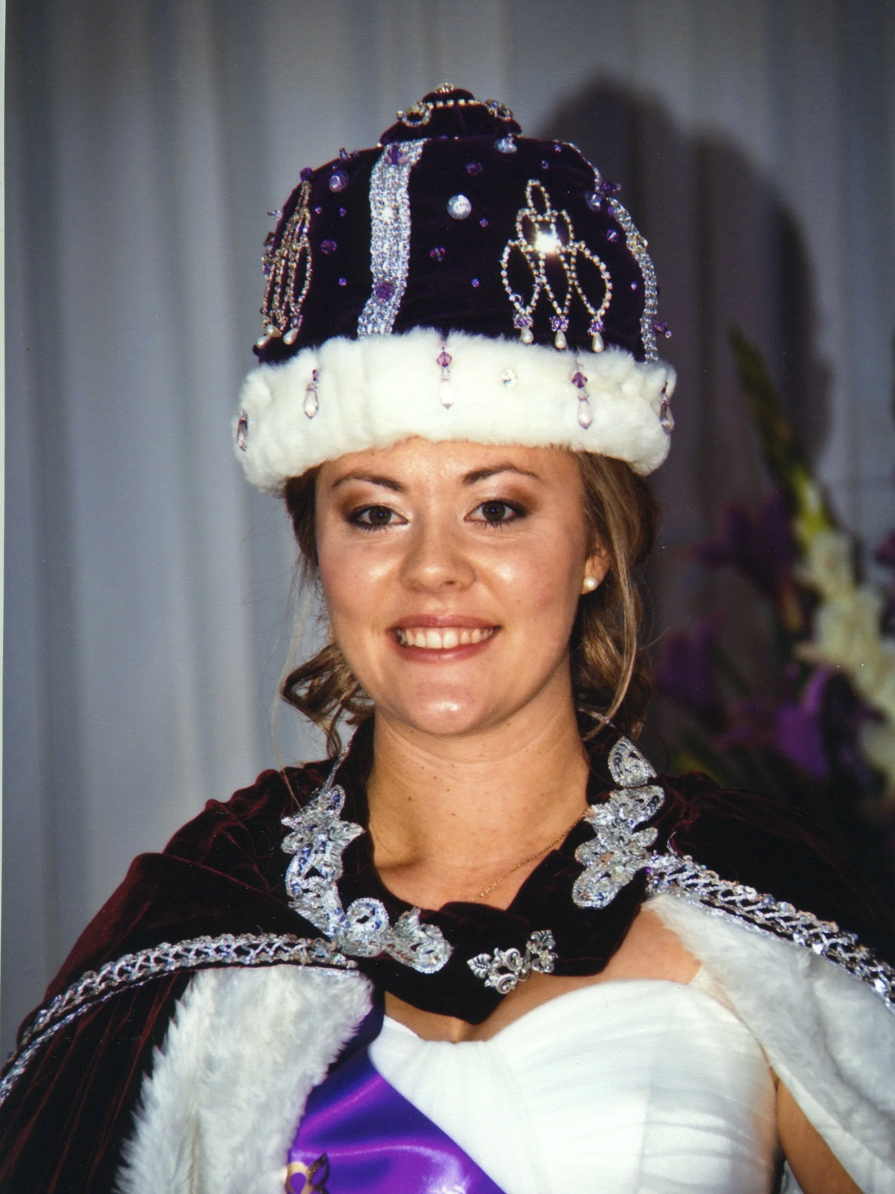 A woman with light brown hair styled in loose waves wearing a dark royal purple velvet crown adorned with silver and purple embellishments, pearls, and gemstones, dressed in regal attire.