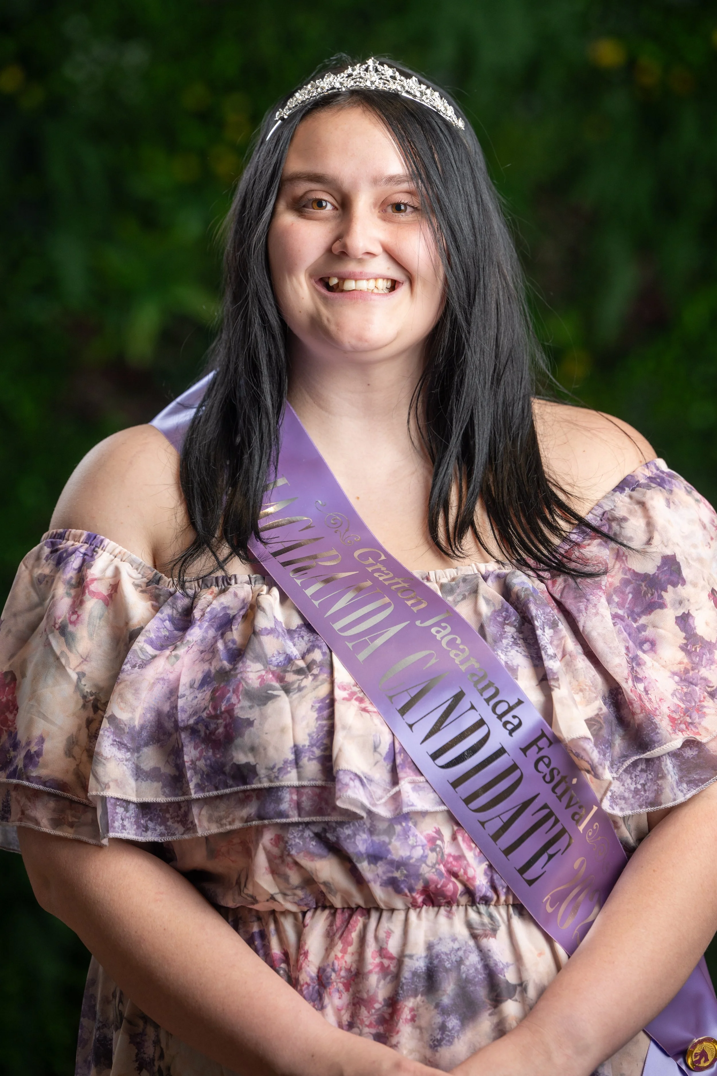 A young woman with dark hair, wearing a tiara, a purple sash that reads "Graduation Jacqueline" and a floral off-the-shoulder dress, smiling outdoors with green foliage in the background.