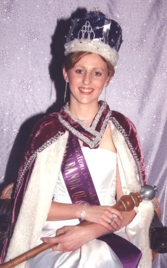 Woman wearing a crown, cape, and sash, holding a scepter, smiling against a sparkly backdrop.