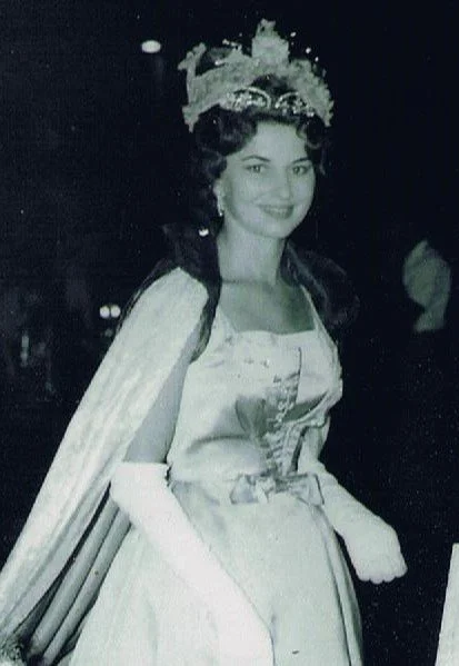 A woman dressed in a historical royal costume with a crown, cape, and gown, smiling at a formal event or celebration.