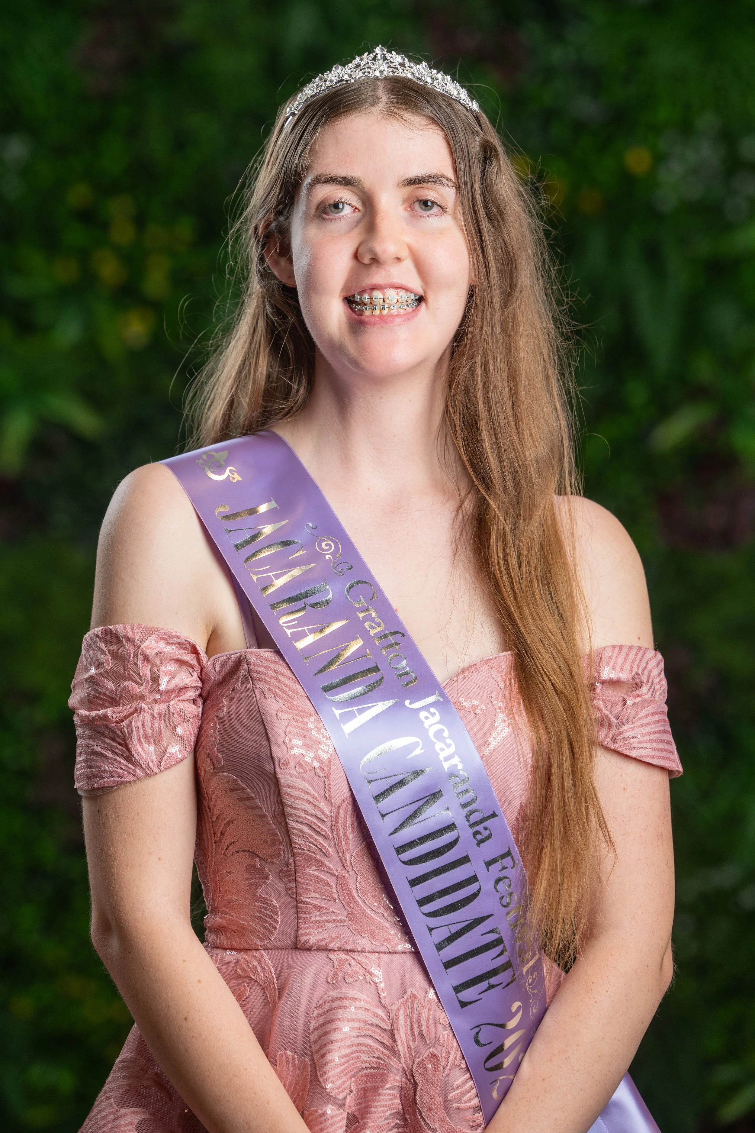Young woman with long red hair wearing a tiara, braces, and a pink dress, standing outdoors against green foliage background, with a lavender sash over her shoulder.